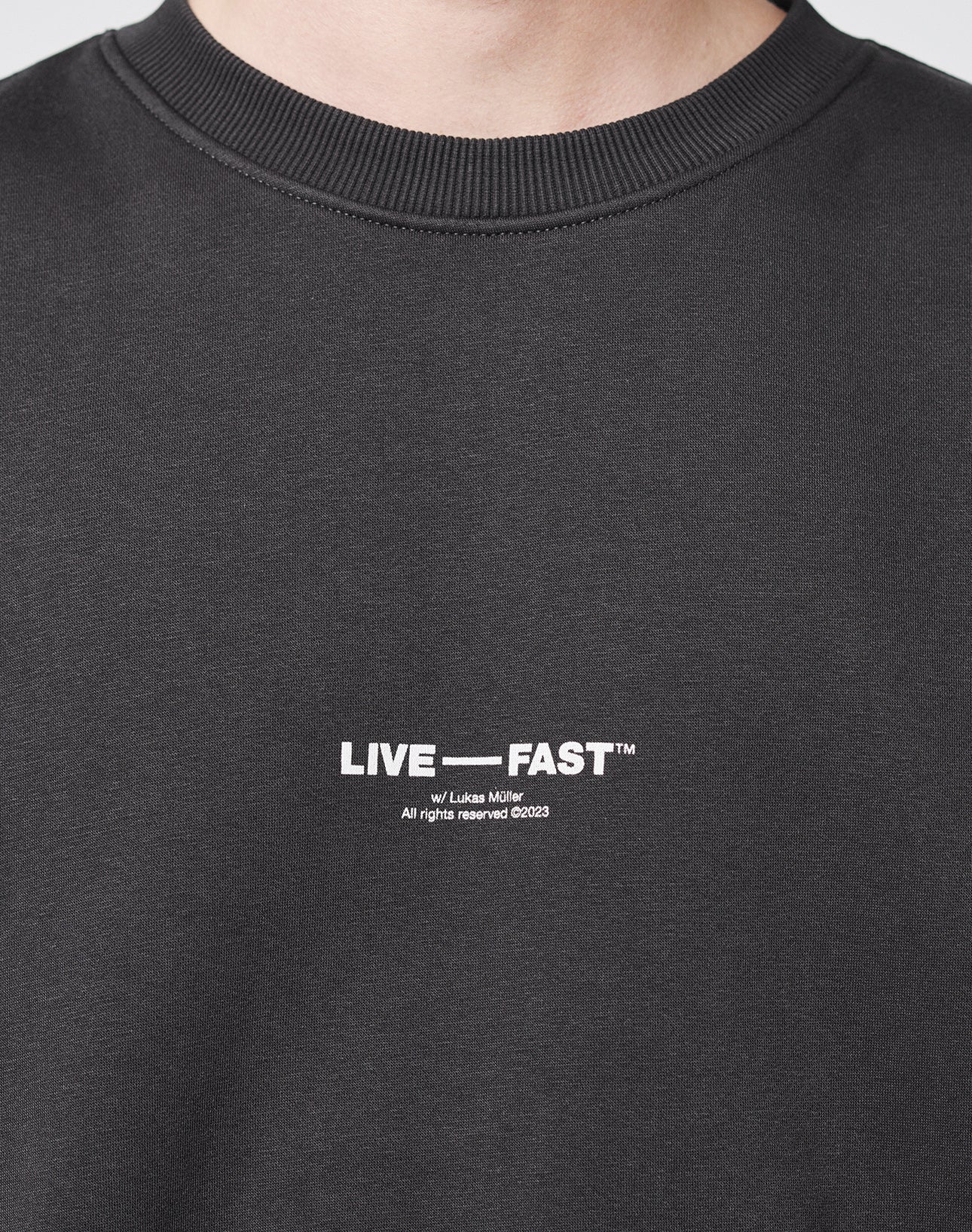 Close-up of a person wearing a dark Tape Sweater by LIVE FAST DIE YOUNG, featuring white LIVE ā FAST⢠text and by Lukas Muller below. Made from a soft cotton-polyester blend. āAll rights reserved Ā©2023ā is printed on the chest.