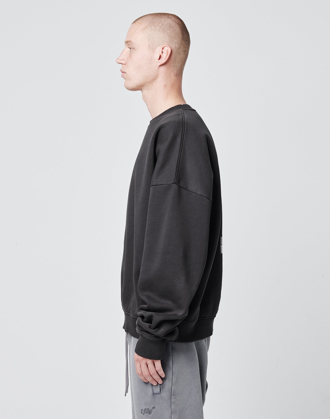 A person with a buzz cut wears the LIVE FAST DIE YOUNG Tape Sweater, an oversize fit dark sweatshirt with cuffed sleeves and hem, paired with light gray pants, standing in profile against a plain light gray background.