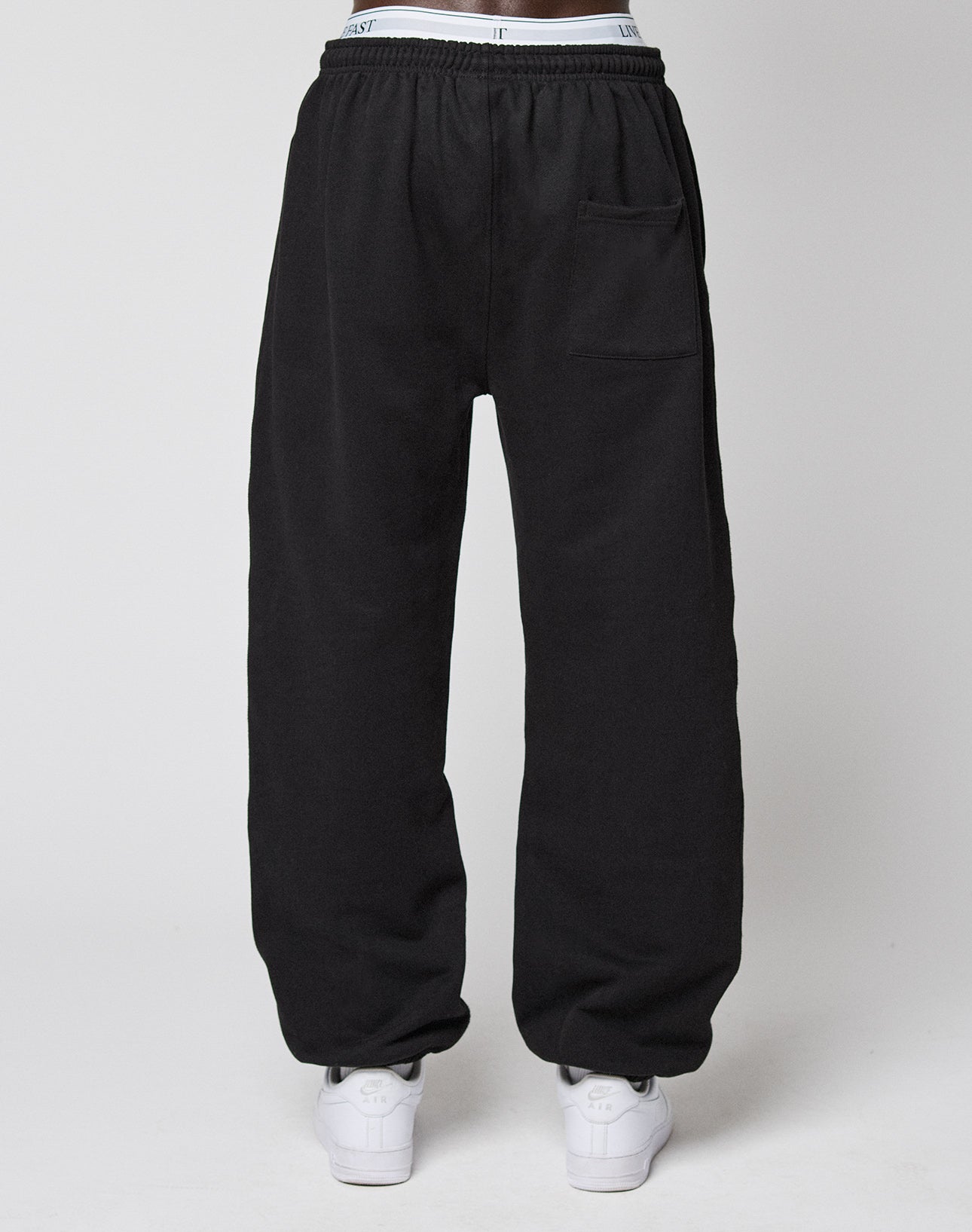A person wearing LIVE FAST DIE YOUNG Wide Cuff Jogger in black, featuring an elastic waistband and a right thigh pocket, paired with white sneakers, stands against a plain white background.