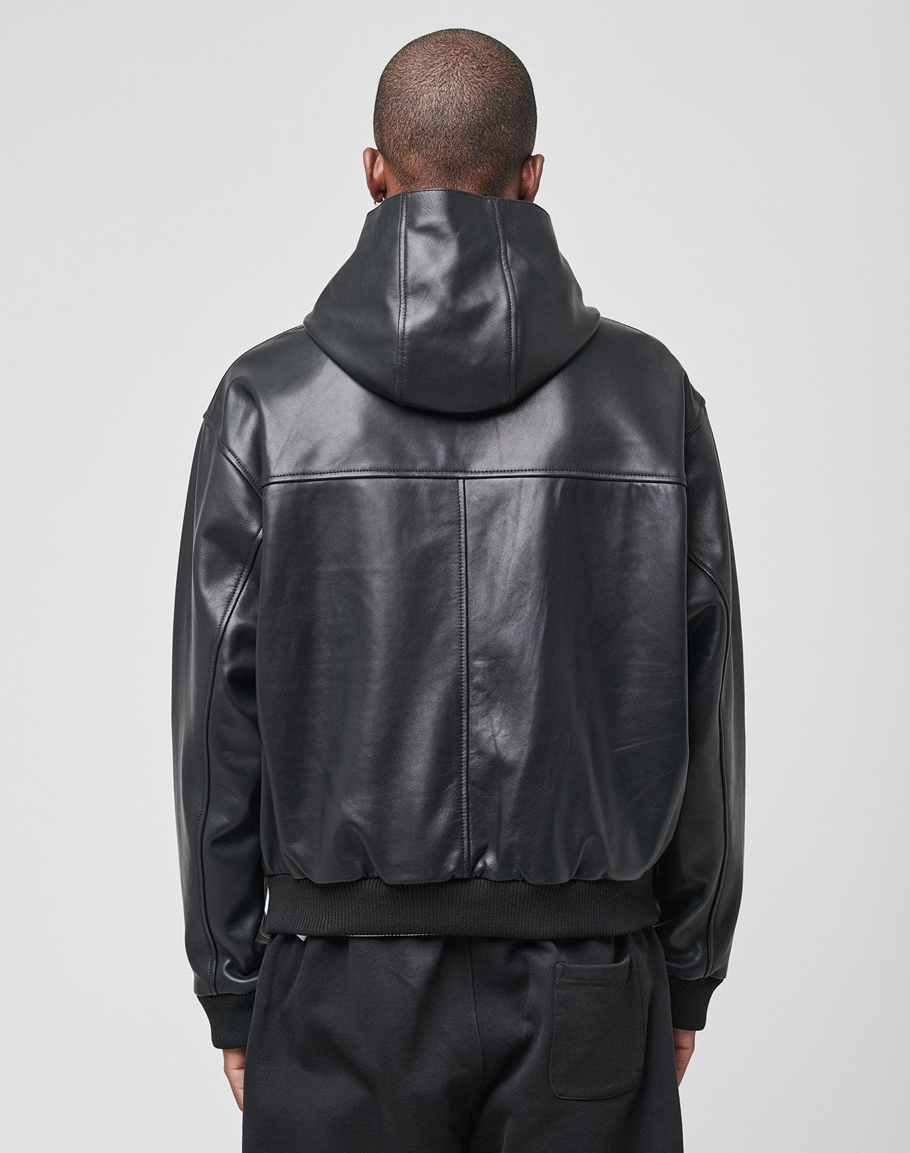 A person with a shaved head is seen from behind, wearing an oversize fit black LIVE FAST DIE YOUNG Leather Hooded Jacket and black pants against a plain light gray background.