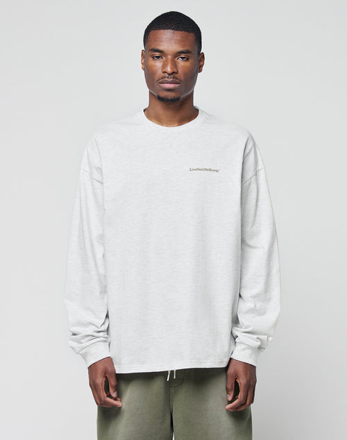 A man with short hair wears the LIVE FAST DIE YOUNG Basic Longsleeve, a light gray, logo-print, oversize sweatshirt, paired with olive green pants, standing against a plain light background.
