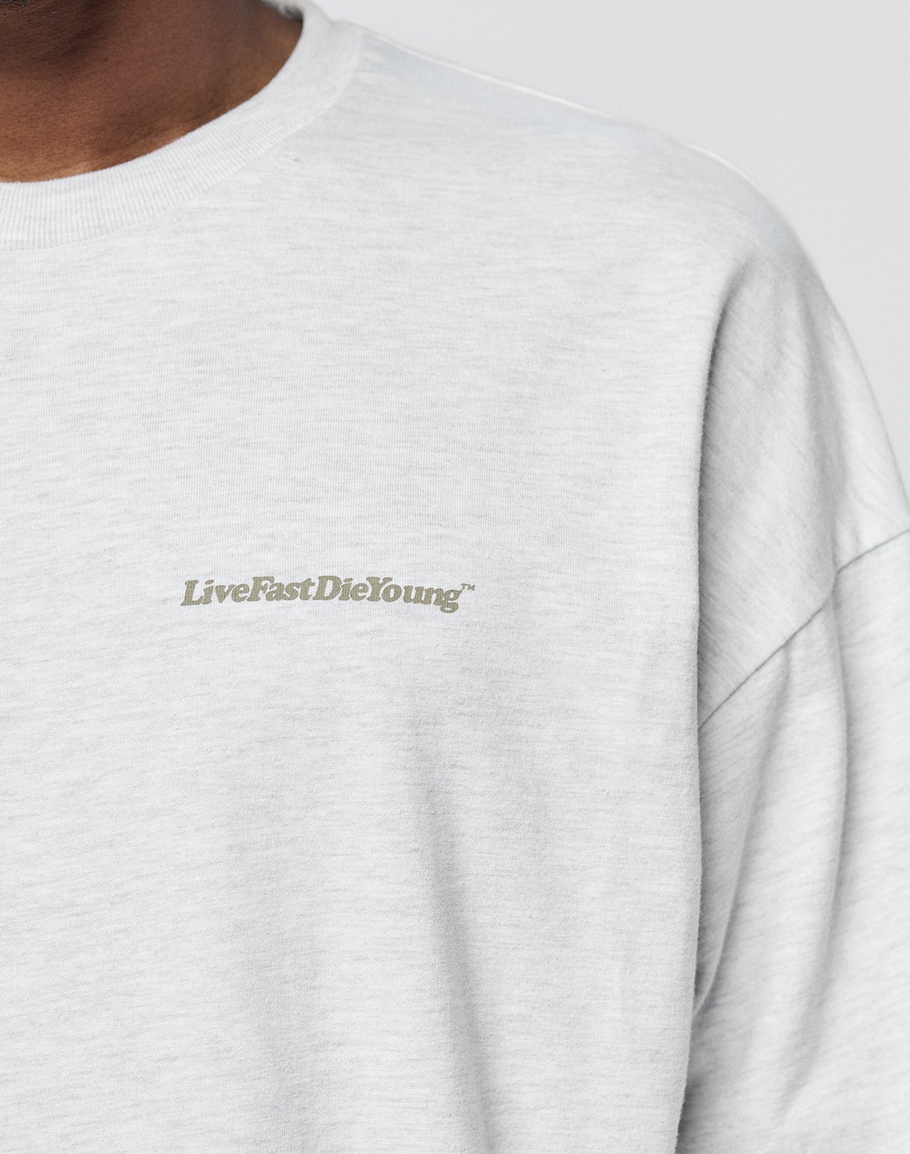 A close-up of someone wearing the LIVE FAST DIE YOUNG Basic Longsleeve, an oversize fit light gray sweatshirt crafted from 100% Baumwolle, with a subtle chest logo-print in small gray āLiveFastDieYoungā capitals. Face not fully shown.