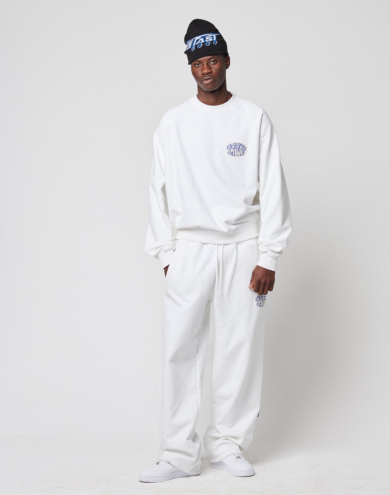 A man stands against a plain background wearing the LFDY Radiator Sweater by LIVE FAST DIE YOUNG, an oversize fit white sweatshirt in Baumwolle Polyester, paired with matching sweatpants, white sneakers, and a black beanie with blue and white writing.
