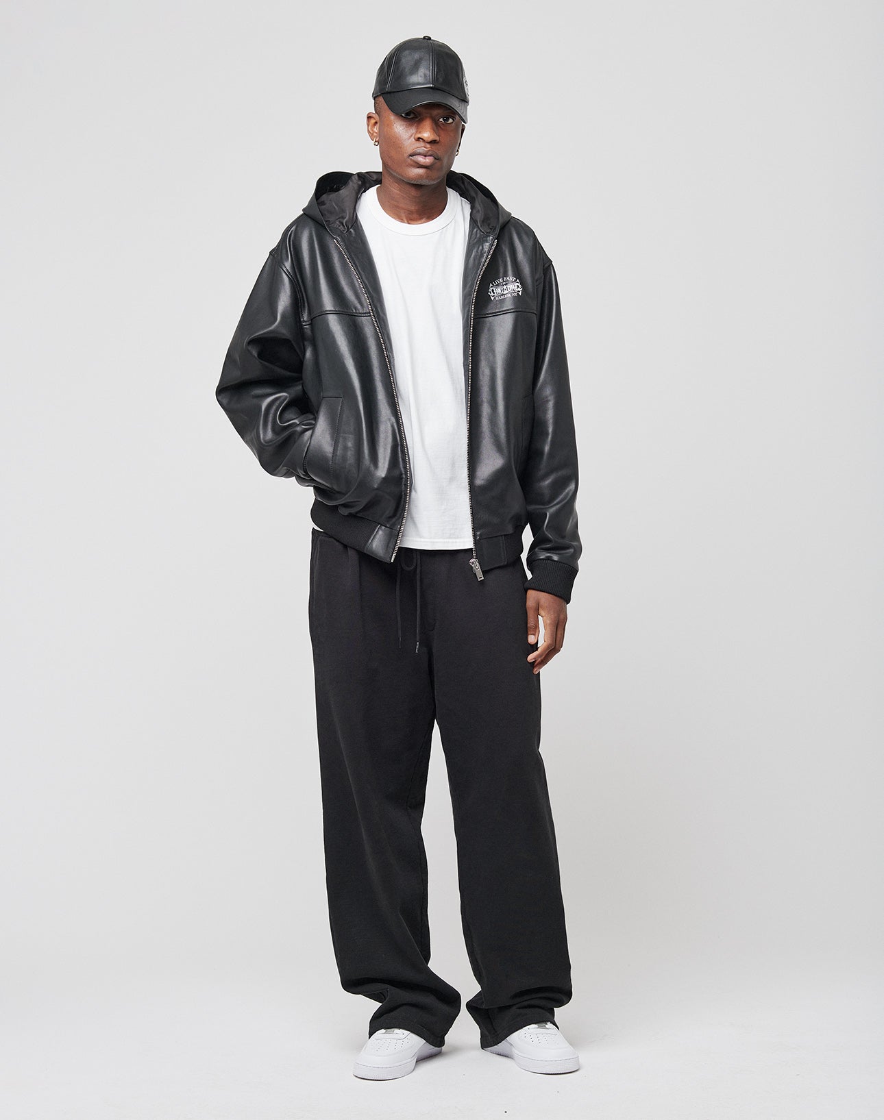 A man in a LIVE FAST DIE YOUNG Leather Hooded Jacket, black pants, white T-shirt, black cap, and white sneakers stands against a plain light background with a neutral expression.