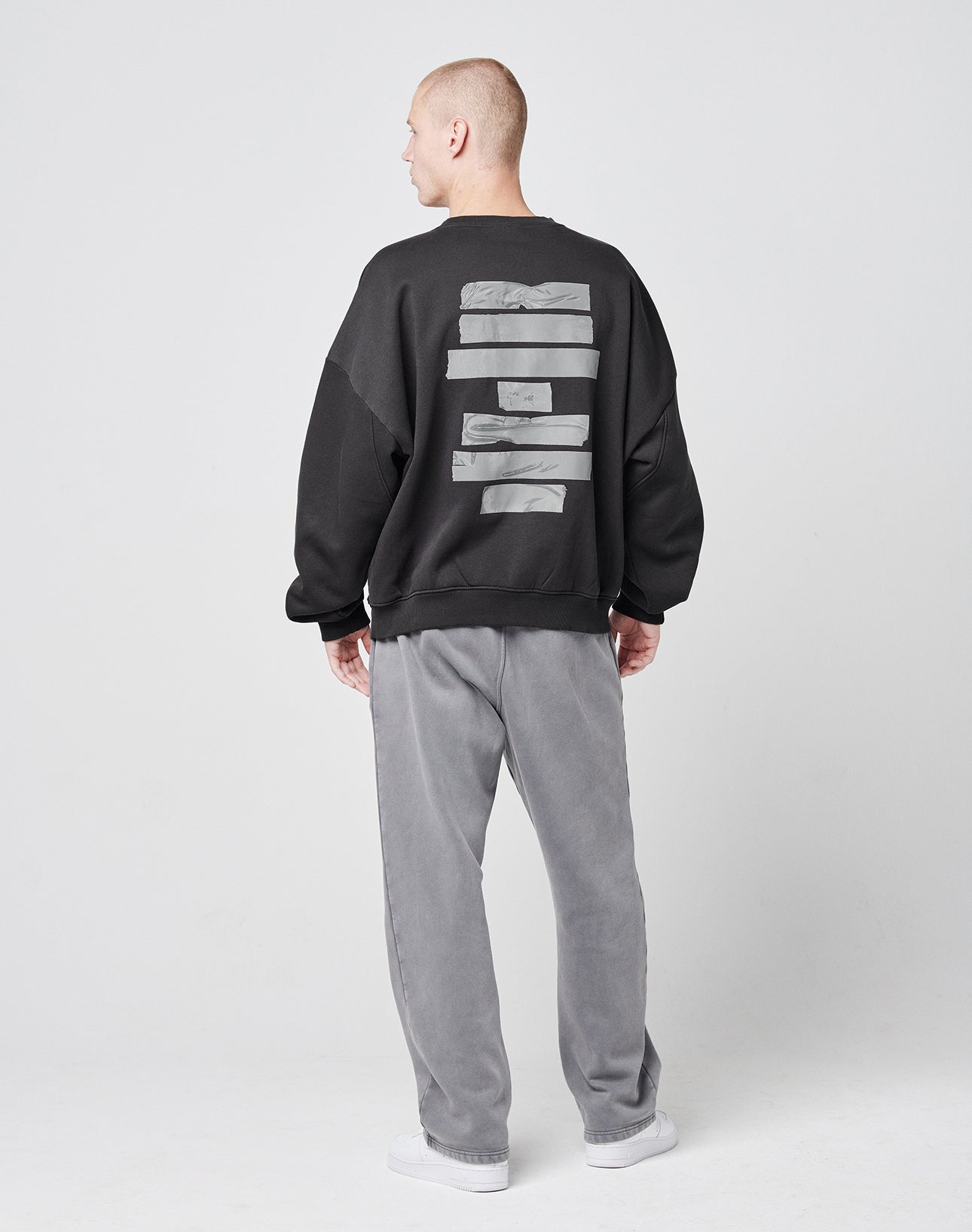 A person stands facing away, wearing the LIVE FAST DIE YOUNG Tape Sweaterāan oversize black Baumwolle and Polyester sweatshirt with gray rectangular designs on the backāpaired with gray pants and white shoes against a plain light background.