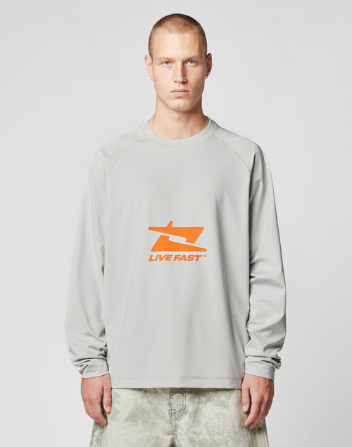 A person with short hair wears the LFDY Sports Longsleeve by LIVE FAST DIE YOUNG, featuring Raglan sleeves and an orange LIVE FAST graphic on light gray, paired with light-colored pants against a plain background.