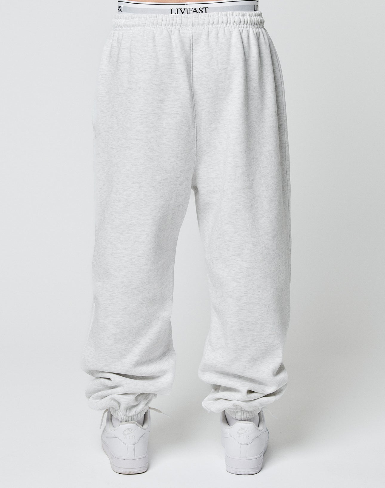Model wears the LIVE FAST DIE YOUNG Basic Loose Jogger in light gray with Elastik-Bund and subtle logo, white LIVFAST waistband showing, paired with white sneakers, photographed from behind against a plain background.
