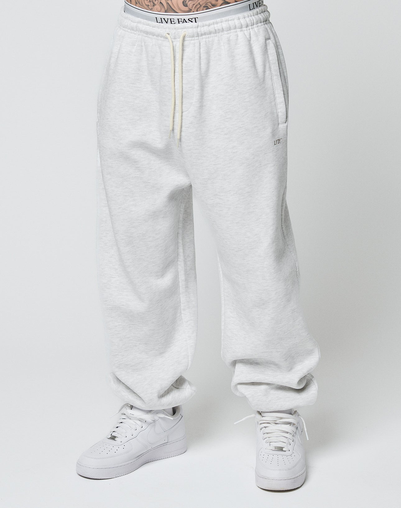 Someone models the LIVE FAST DIE YOUNG Basic Loose Jogger in light gray with a Loose Fit and elastic waistband featuring LIVE FAST text, paired with white sneakers, standing against a plain white background.