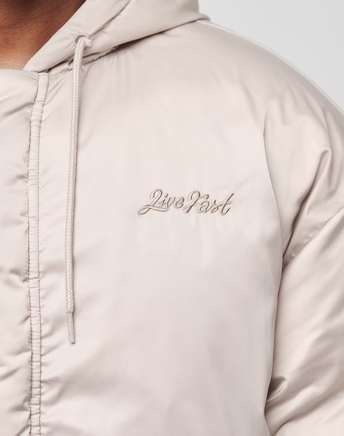 Close-up of a person wearing the LFDY Winter Bomber Jacket by LIVE FAST DIE YOUNG, a light beige oversized jacket with drawstring hood (Kapuze) and Live Fast cursive embroidery on the left chest. Partially visible zipper adds detail.