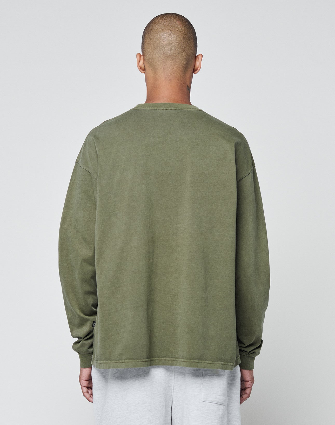 A person with a shaved head faces away from the camera, wearing the LIVE FAST DIE YOUNG Basic Longsleeve in olive green (100% Baumwolle, oversize fit) and light gray pants, posed against a plain light gray background.