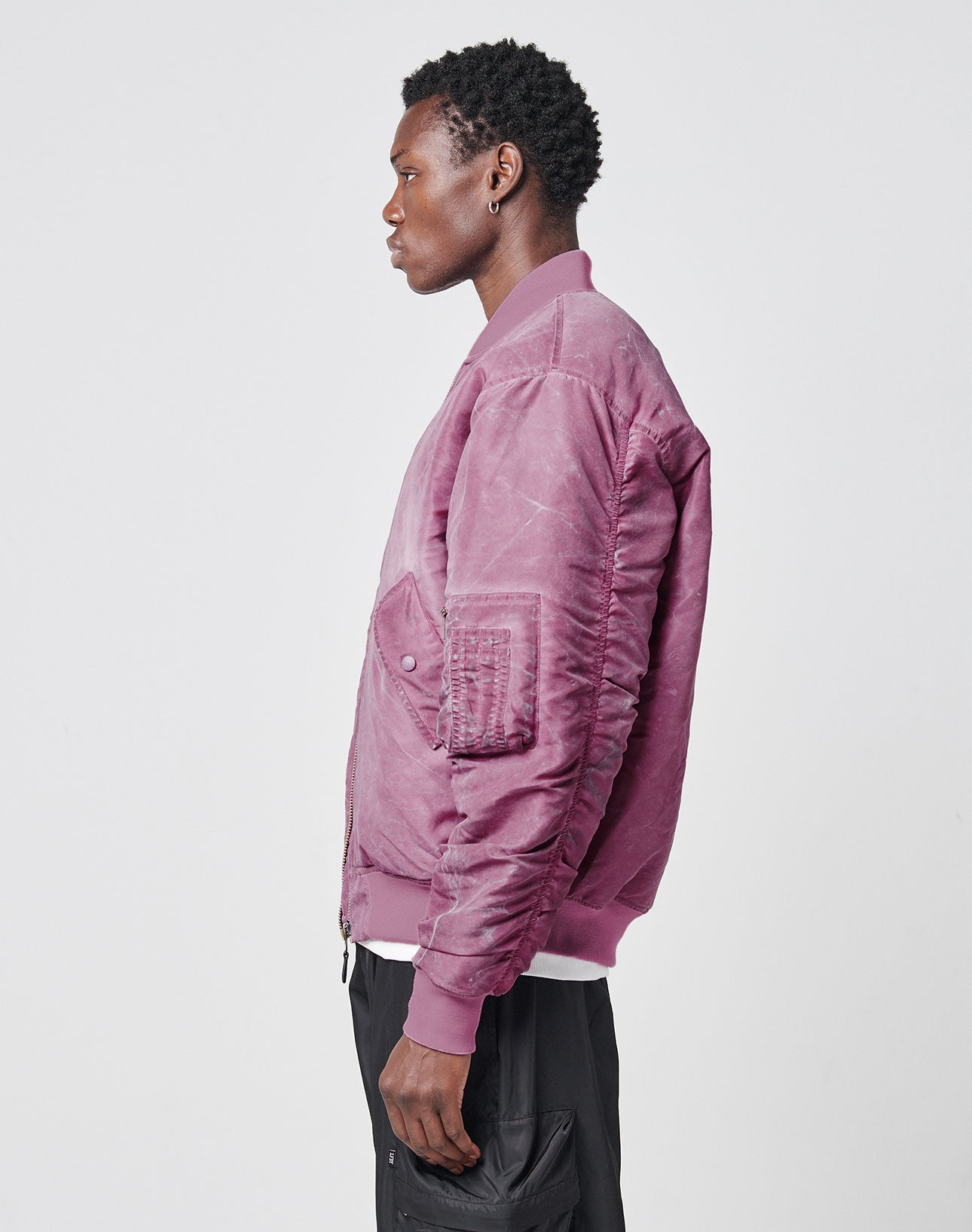 A person with short curly hair wears the LIVE FAST DIE YOUNG Washed Bomber Jacket in mauve, featuring a subtle stickpatch, over a white shirt and black pants, standing in profile against a plain light background.