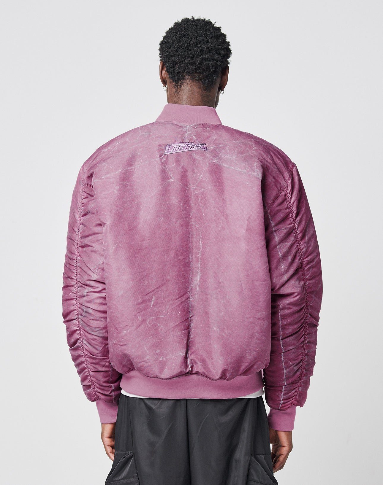 A person faces away, wearing the LIVE FAST DIE YOUNG Washed Bomber Jacketāan oversized mauve piece with ruched sleeves, a small embroidered logo, and Stickpatch on the upper backāpaired with black pants against a plain background.