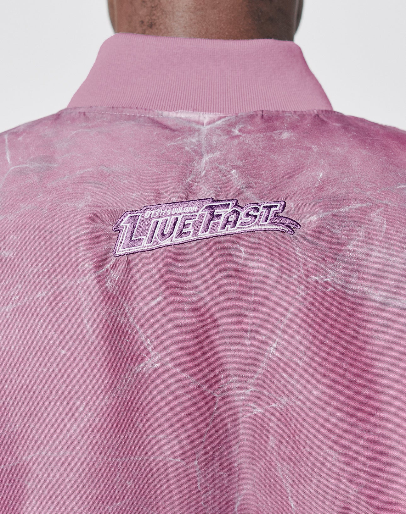 A close-up of the Washed Bomber Jacket by LIVE FAST DIE YOUNG shows pink marbled fabric and LIVE FAST embroidered in white and purple near the collar, finished with a subtle vintage wash for a sporty retro look.