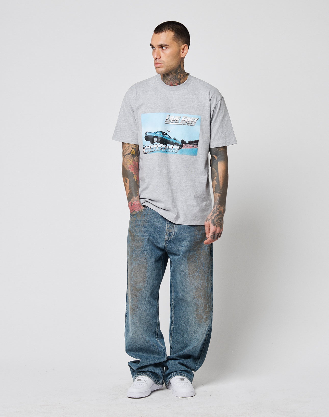 A tattooed man faces left, wearing the LFDY Crash Kid Tee by LIVE FAST DIE YOUNG—a gray graphic regular fit T-shirt with a car and text—paired with loose blue jeans and white sneakers, against a plain light background.