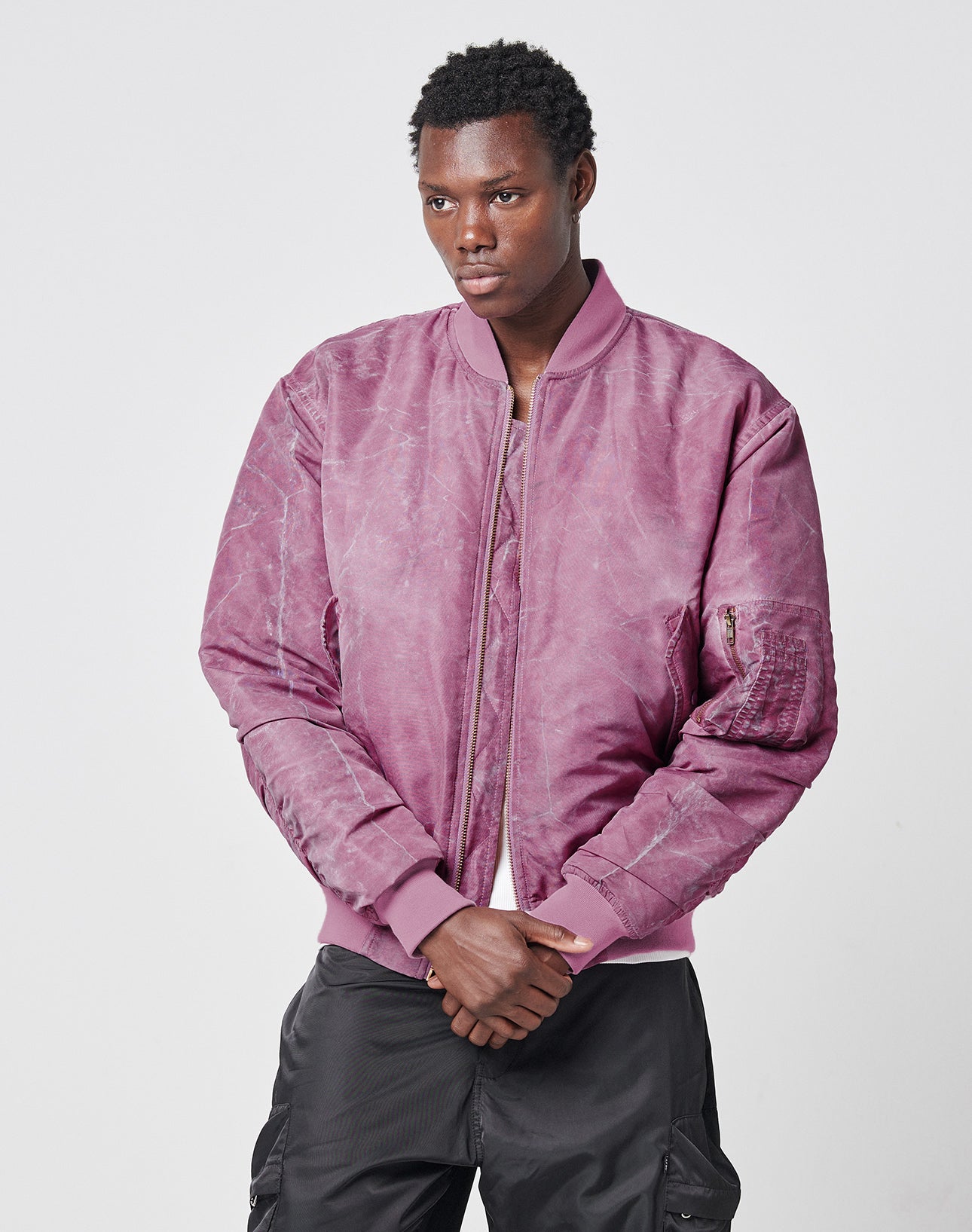 A person wears the LIVE FAST DIE YOUNG Washed Bomber Jacket in mauve with black cargo pants, standing against a plain light gray background and looking slightly to the side with hands clasped in front.