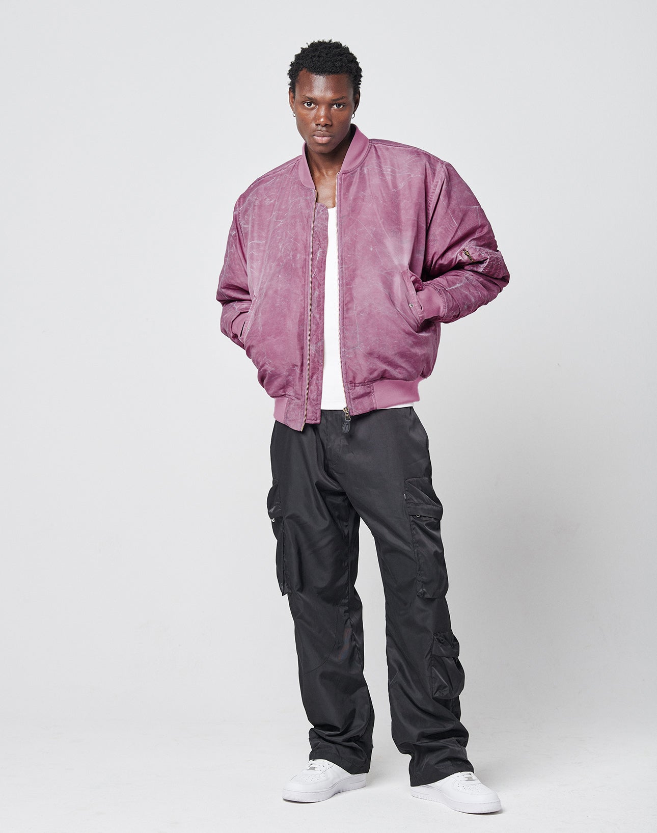 A man stands against a plain white background wearing the LIVE FAST DIE YOUNG Washed Bomber Jacket, a white t-shirt, black cargo pants, and white sneakers, with his hands tucked into his jacket pockets.