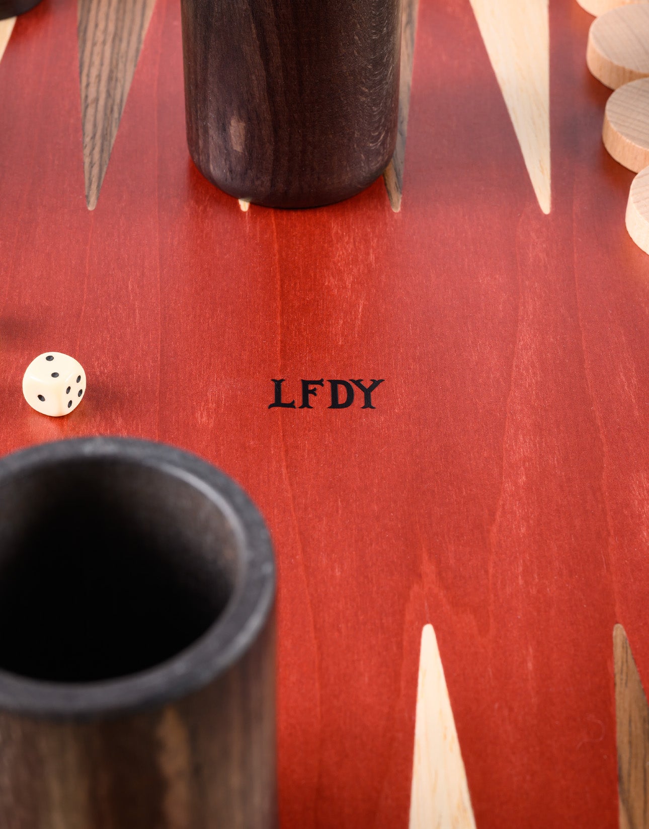 A close-up of the LFDY Backgammon board by LIVE FAST DIE YOUNG shows red and beige triangles, dark Walnuss Holz dice cups, a die with five dots, beige pieces, and "LFDY" in the center. Handcrafted in Greece.