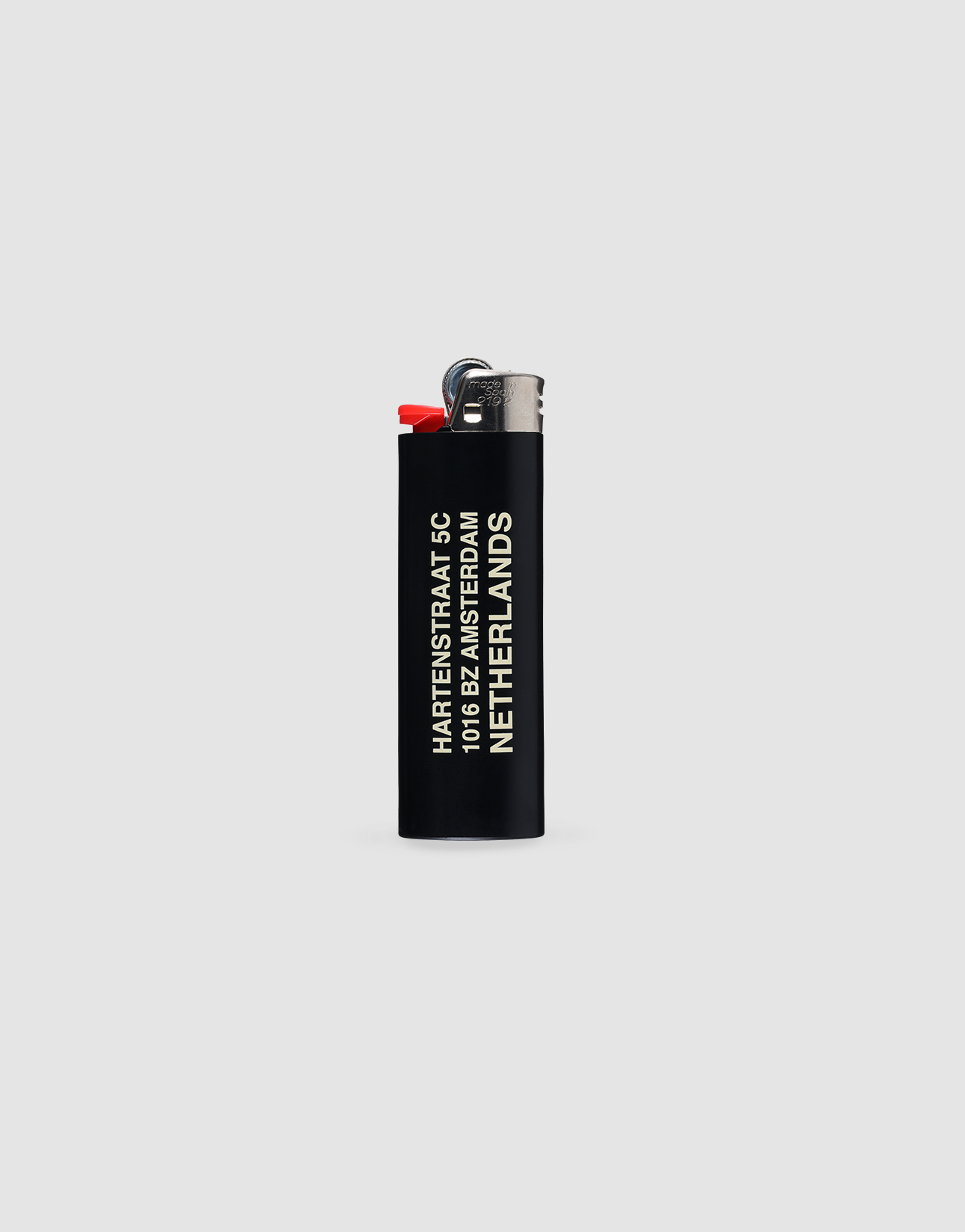 A black LFDY x The Bulldog Lighter by LIVE FAST DIE YOUNG with chrome cap and red ignition stands upright. āHartenstraat 6, 1016 BE Amsterdam, Netherlandsā is printed in white. Up to 3000 ignitions. Light gray background.