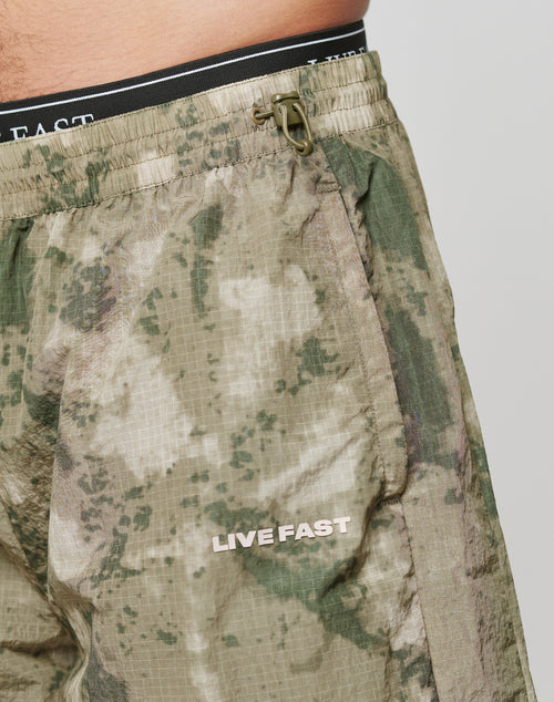 Close-up of a person wearing LIVE FAST DIE YOUNG Live Fast Nylon Shorts in green camo with oversized fit, black waistband with white text, side pockets, LIVE FAST Silikon-Print on the leg, and drawstring with plastic fastener.