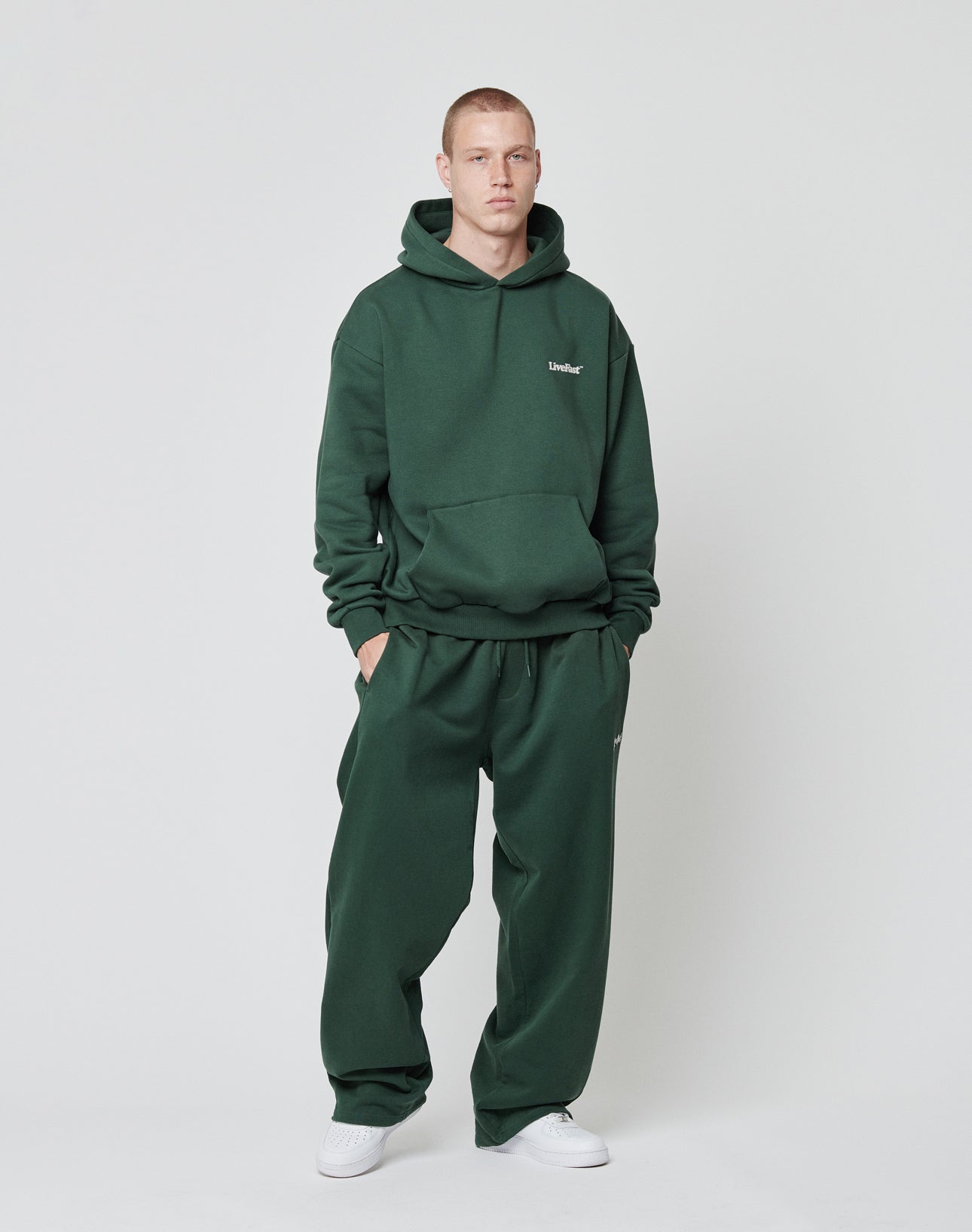 A person stands against a plain background wearing LIVE FAST DIE YOUNGs Basic Wide Jogger in dark green with a matching hoodie and an elastic waistband, paired with white sneakers, hand in pocket and a neutral expression.