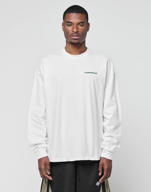 A man faces forward in a LIVE FAST DIE YOUNG Basic Longsleeve, an oversized white cotton shirt with a small green logo print on the chest, paired with black shorts featuring tan side panels, against a plain light gray background.