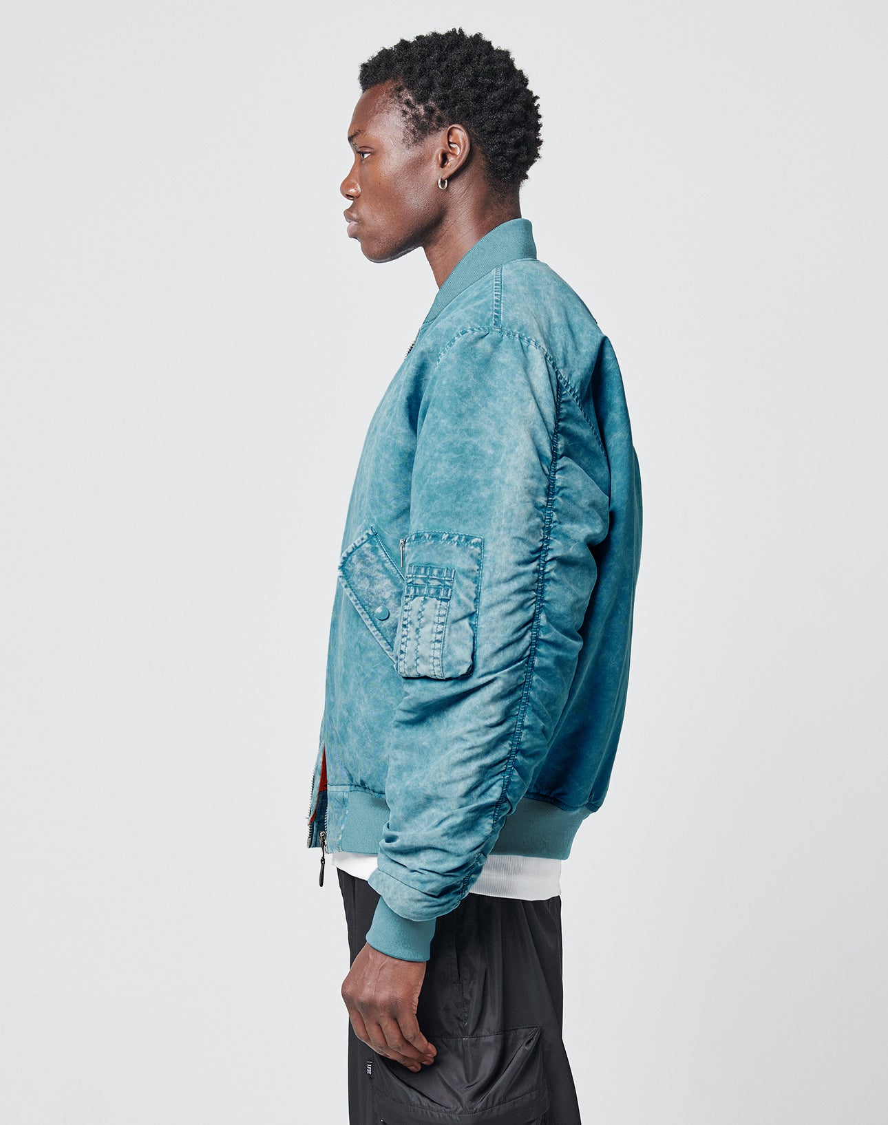 A man with short curly hair stands in profile against a plain background, wearing the LIVE FAST DIE YOUNG Washed Bomber Jacket over a white shirt and black pants.