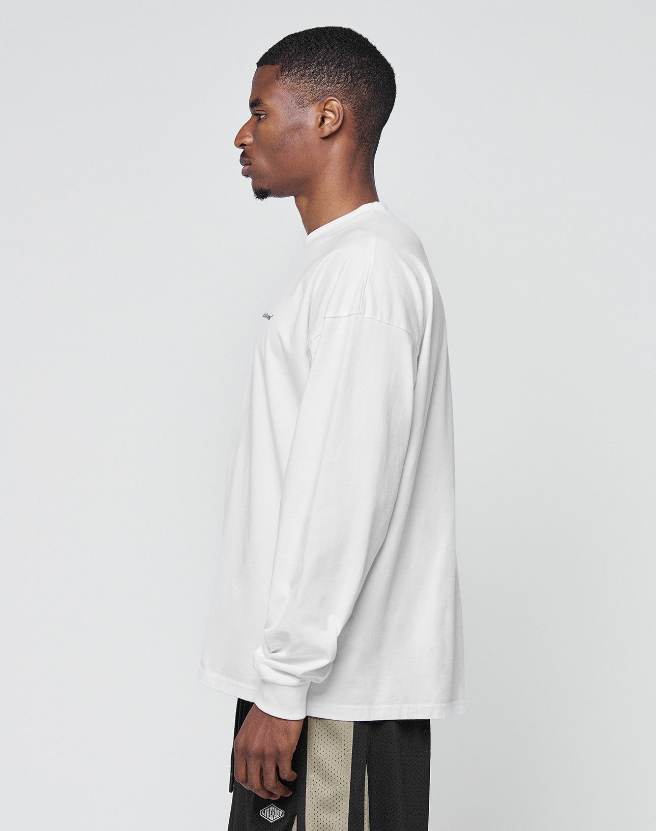 A man with short hair stands in profile against a light background, wearing a LIVE FAST DIE YOUNG Basic Longsleeve in white and black shorts with beige stripes.