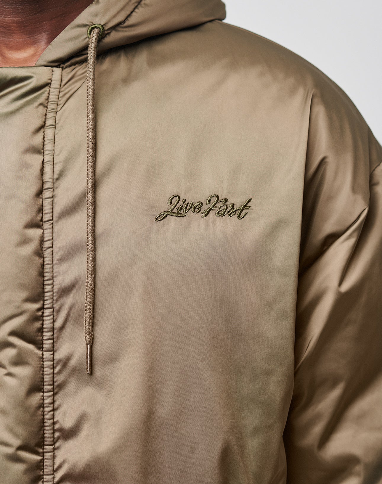 Close-up of a person wearing the LFDY Winter Bomber Jacket by LIVE FAST DIE YOUNG, in light brown satin with an oversize fit, hood with drawstrings, and the Live Past cursive logo embroidered on the left chest.
