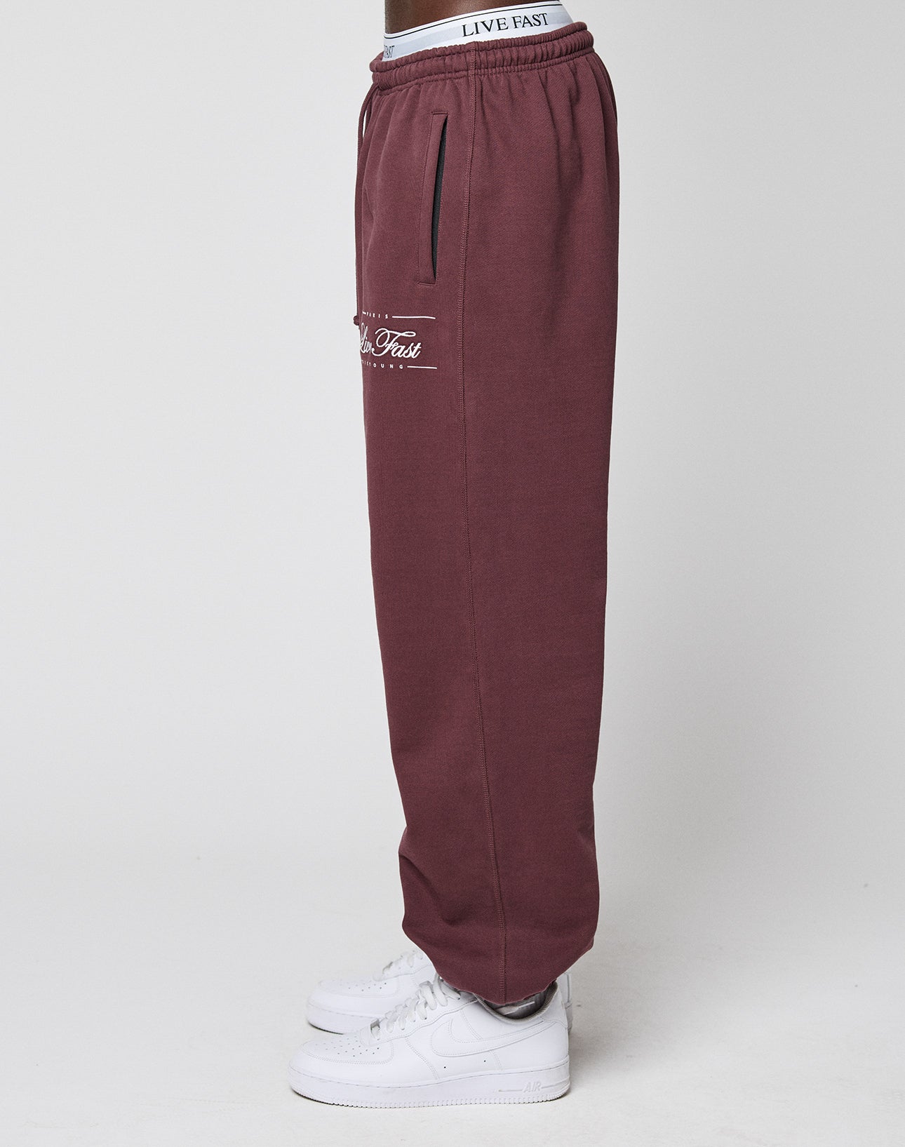 A person wearing LIVE FAST DIE YOUNG Wide Cuff Jogger in maroon, 100% Baumwolle, with "Live Fast" text on the pocket and waistband, pairs them with white sneakers against a plain light background, standing in profile.