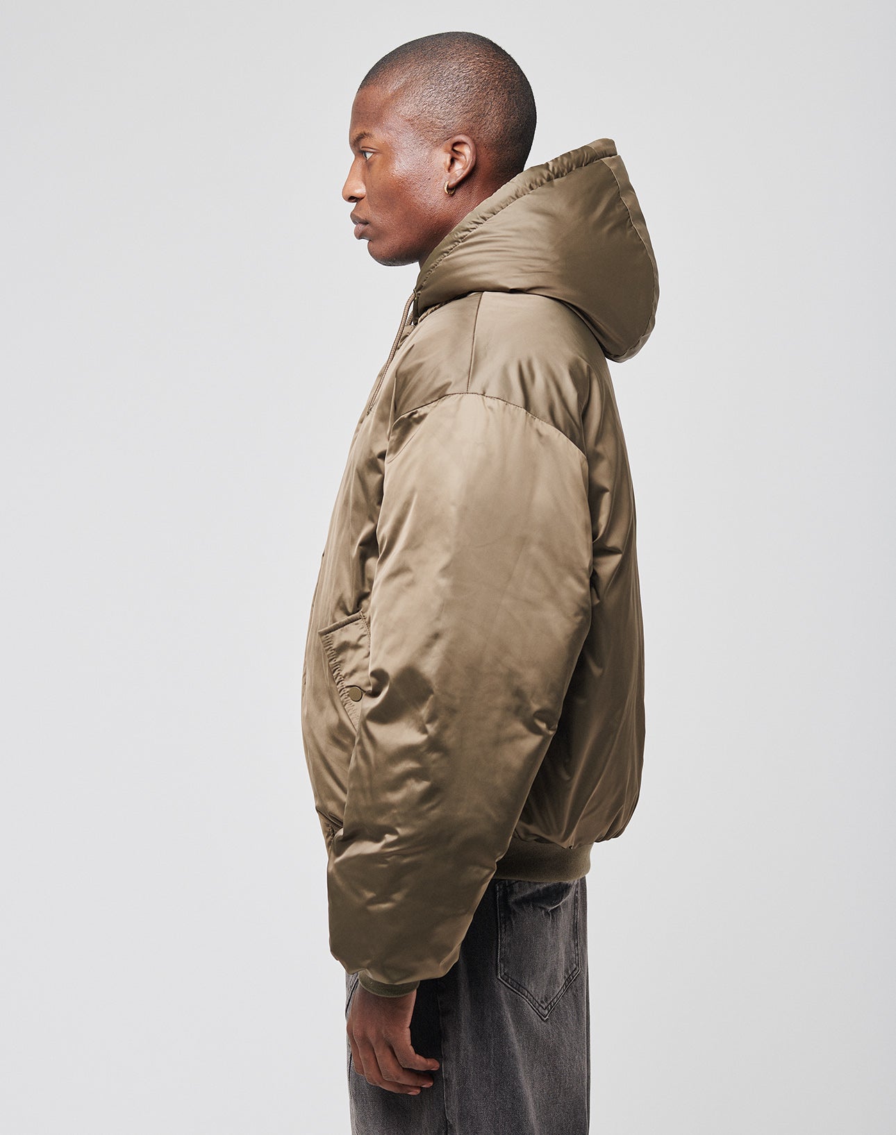 A man stands in profile against a plain light background wearing the LIVE FAST DIE YOUNG LFDY Winter Bomber Jacket in an oversized puffy olive green style with hood and drawstrings, paired with dark jeans.