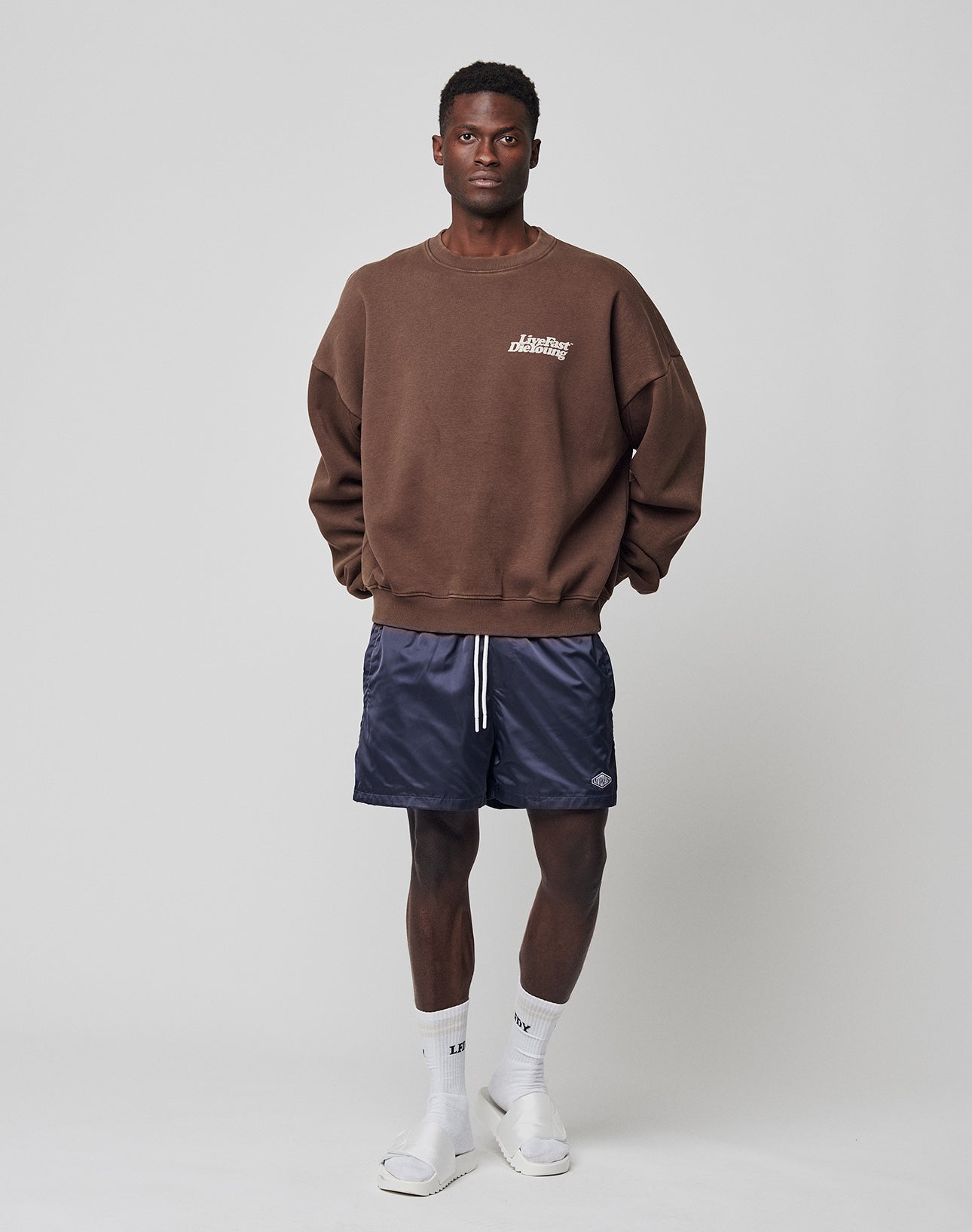 A man faces forward against a light gray background, wearing LIVE FAST DIE YOUNG Basic Satin Shorts in navy, a brown regular fit sweatshirt with a logo patch, white crew socks, and white slide sandals. His hands are in his sweatshirt pockets.