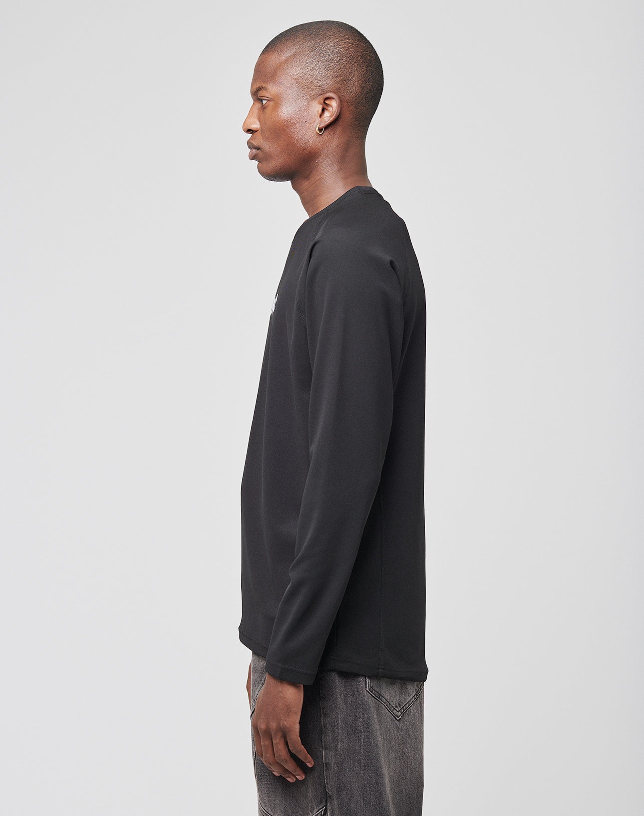A man in profile faces left, wearing a LIVE FAST DIE YOUNG Live Fast Sports Longsleeve (100% Polyester) and gray jeans, standing against a plain light gray background.