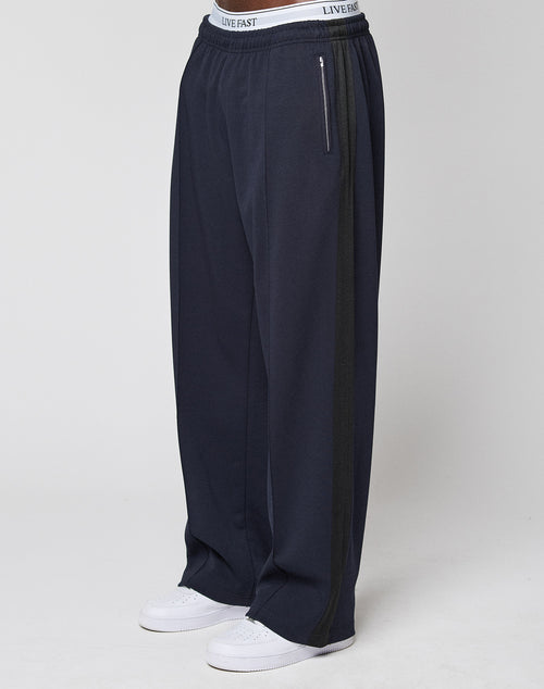 Wearing the LFDY Striped Jogger by LIVE FAST DIE YOUNG, featuring a loose fit, navy blue color, dark Fischgräten-Tape side stripe, and a white LIVE FAST waistband, paired with white sneakers against a plain light background.