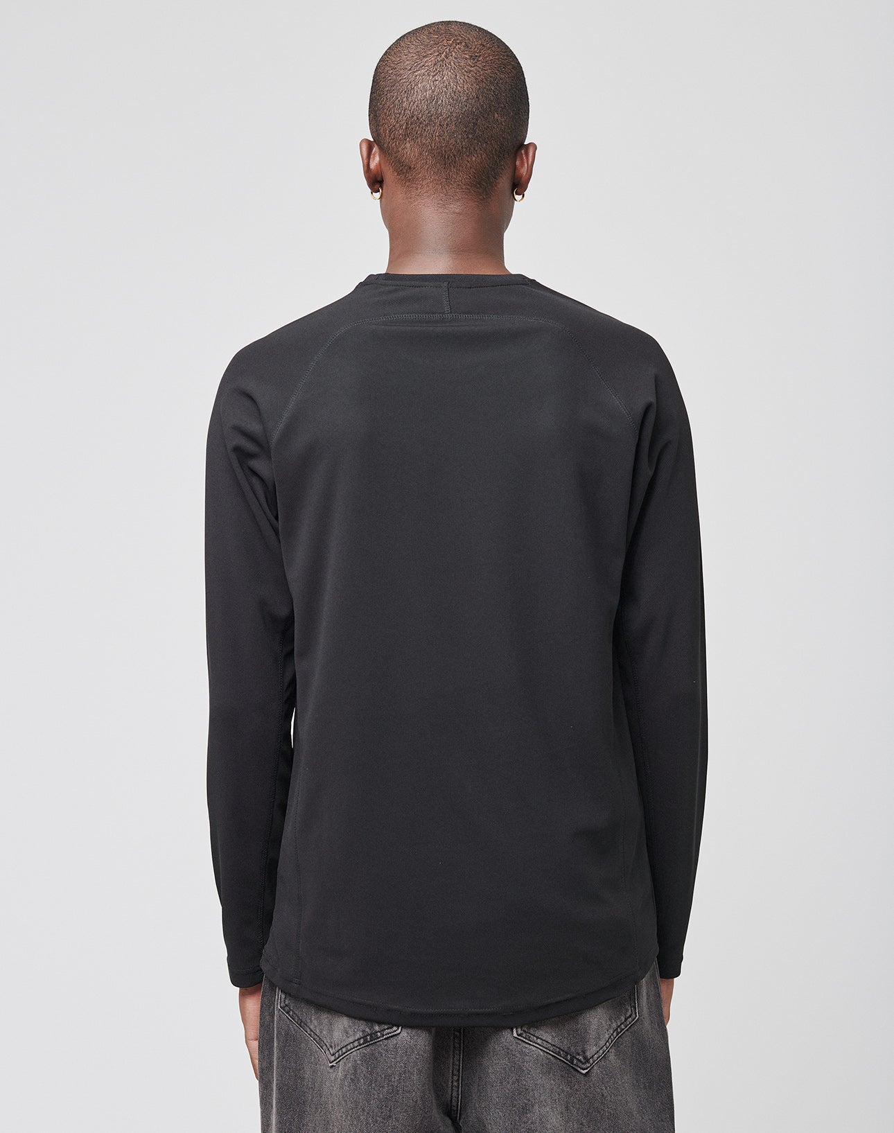 A person with a shaved head is shown from behind, wearing a LIVE FAST DIE YOUNG Live Fast Sports Longsleeve and faded black jeans against a plain light gray background.