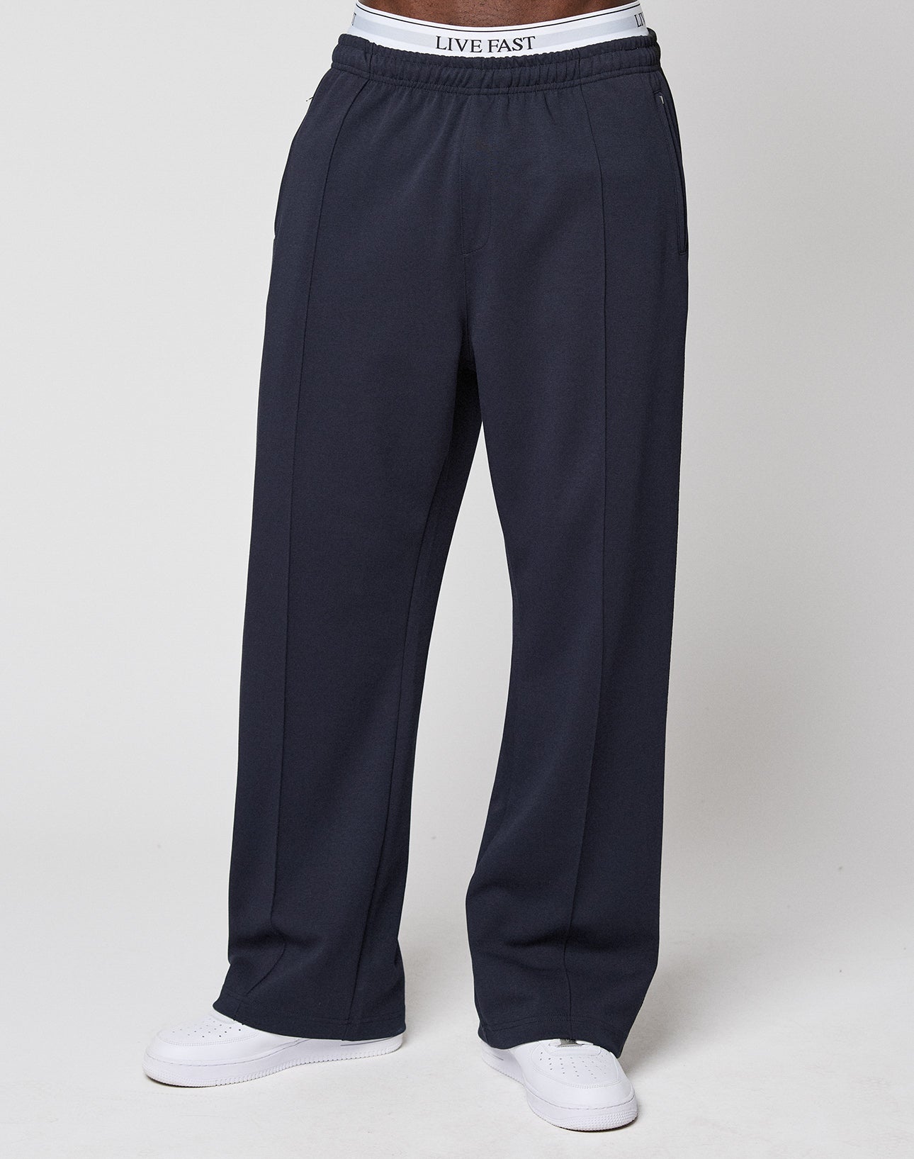 A person wearing loose-fitting dark navy LFDY Striped Jogger sweatpants by LIVE FAST DIE YOUNG, paired with white sneakers and a visible white LIVE FAST waistband, stands against a plain light background.