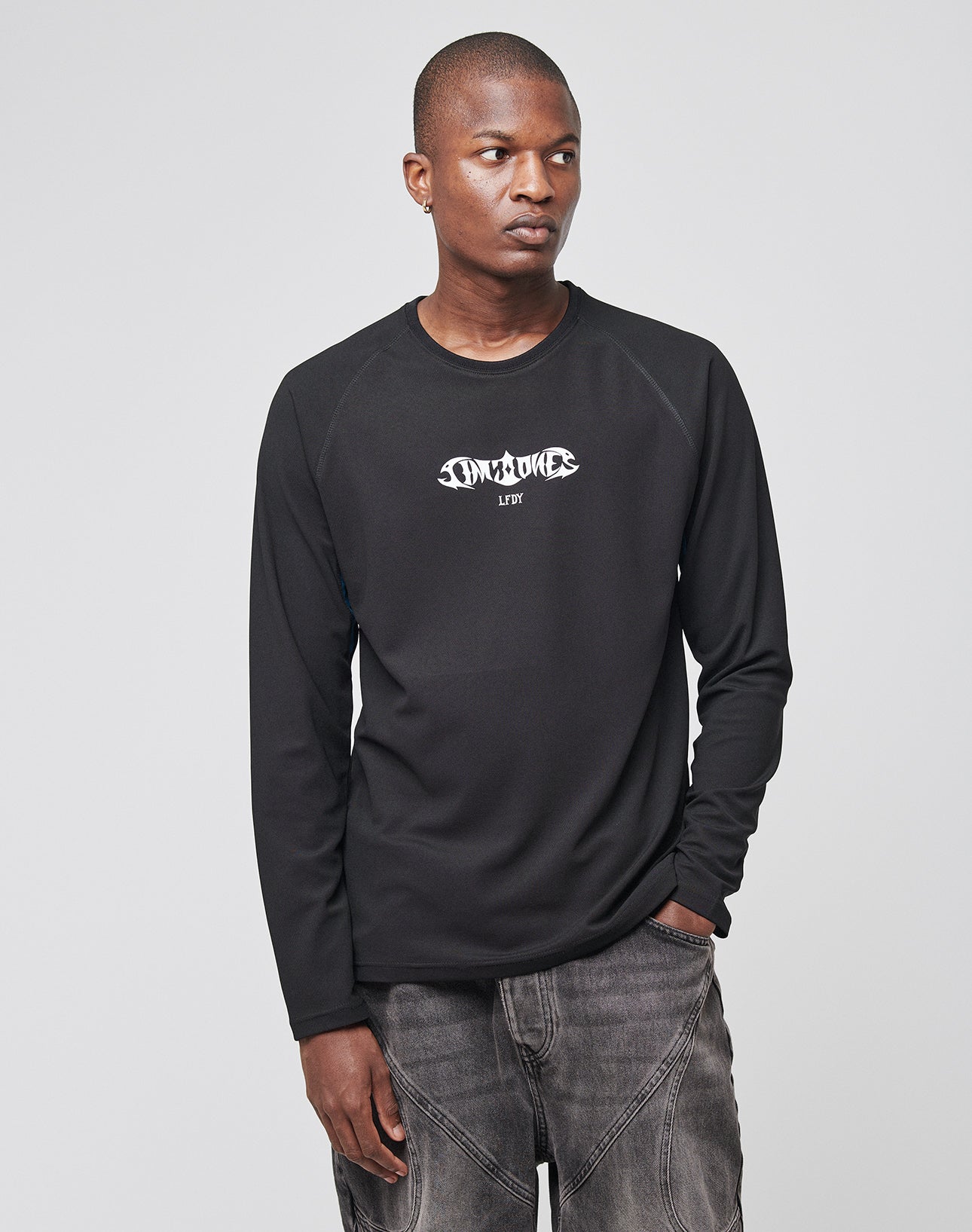 A person in a Live Fast Sports Longsleeve by LIVE FAST DIE YOUNG, featuring a stylized white logo and gray jeans, stands against a plain light gray background, looking to the side with one hand in their pocket. Model: LF2310-14898.