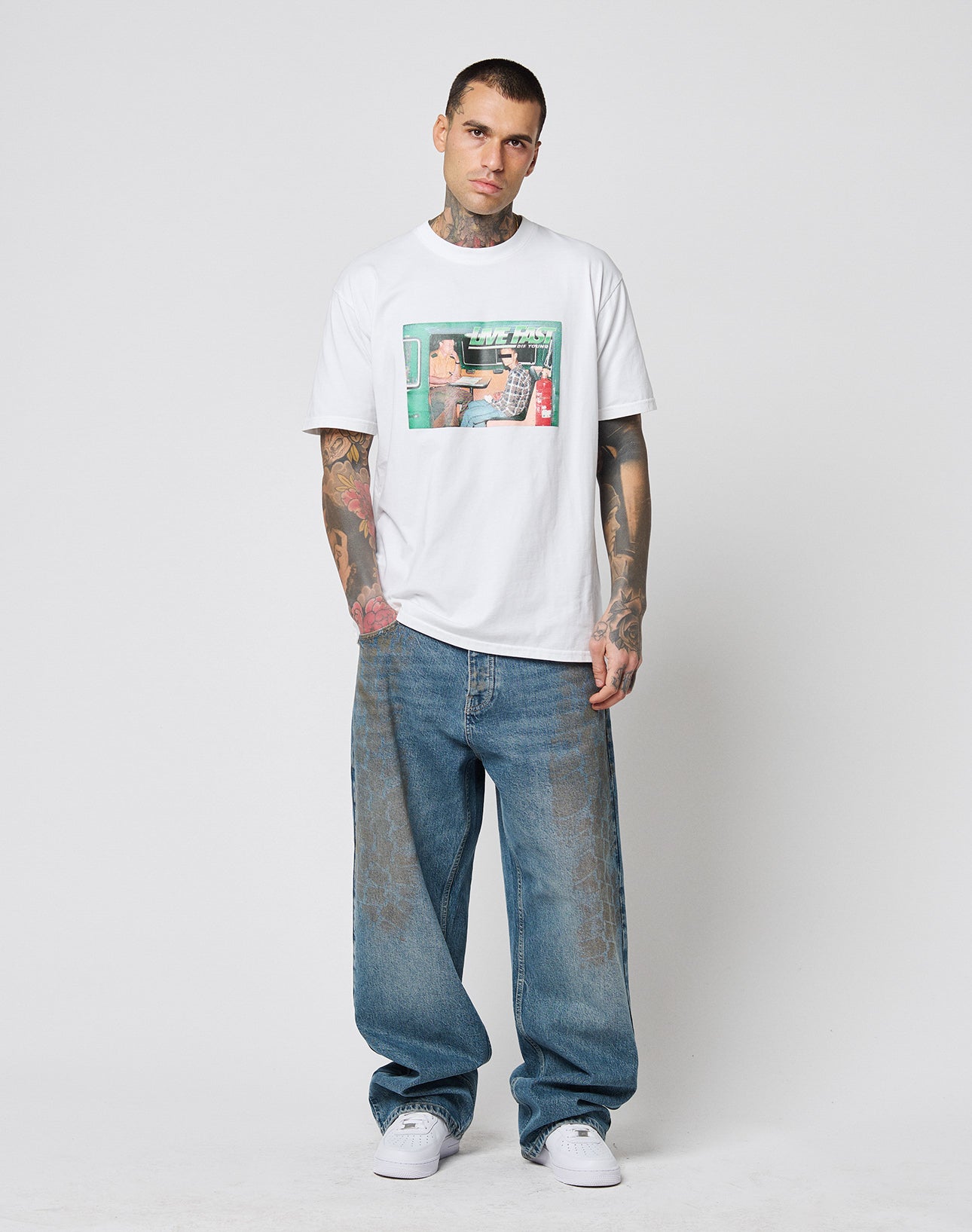 A tattooed man faces the camera in a white LIVE FAST DIE YOUNG LFDY Sixers Tee (Regular Fit, 100% Baumwolle), loose blue jeans, and white sneakers against a gray background. Hands relaxed by his sides. Waschhinweise included.