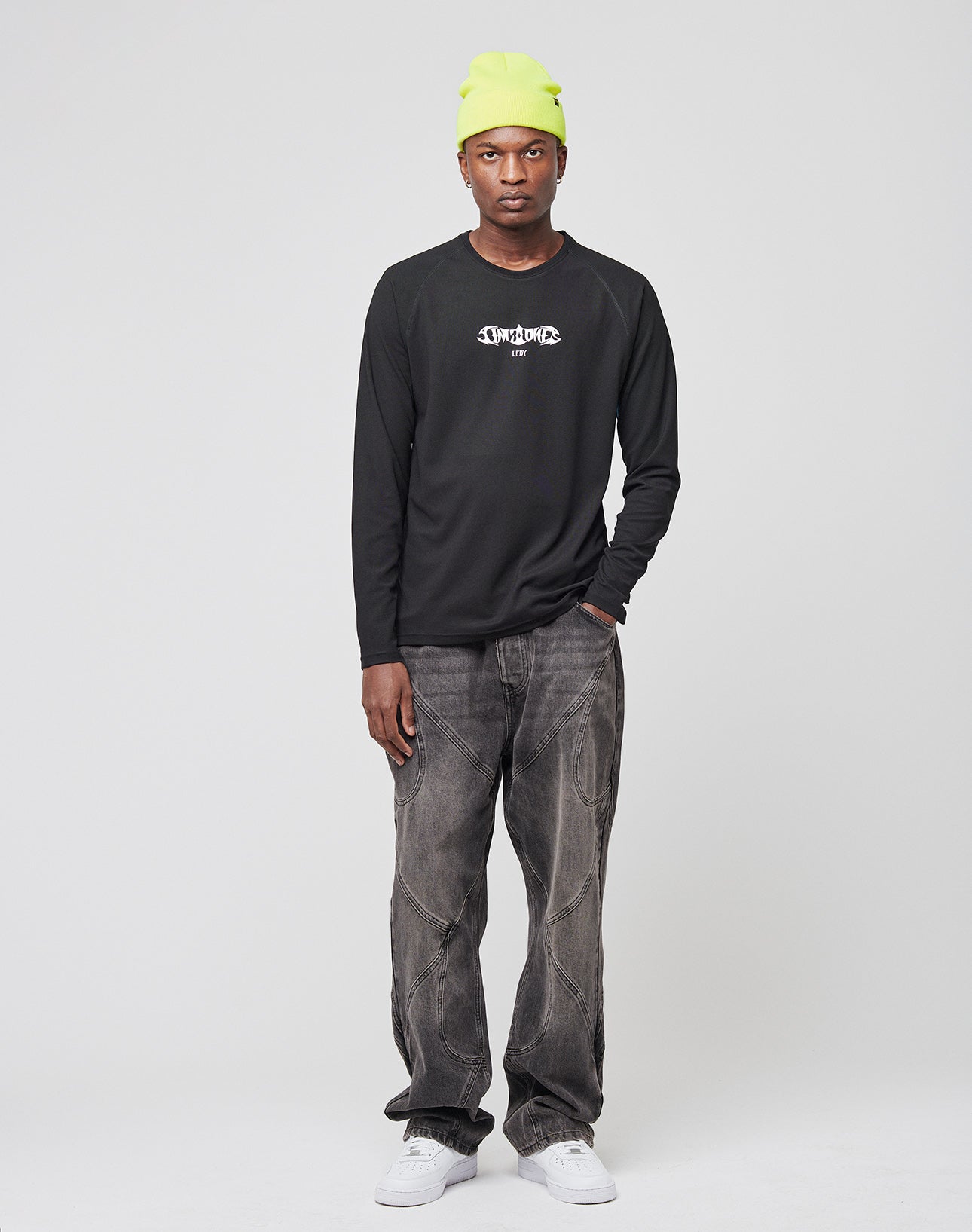 A man stands against a white background wearing a neon yellow beanie, the LIVE FAST DIE YOUNG Live Fast Sports Longsleeve in black with a white graphic, faded baggy jeans, and white sneakers. His expression is neutral and his hands are relaxed by his sides.