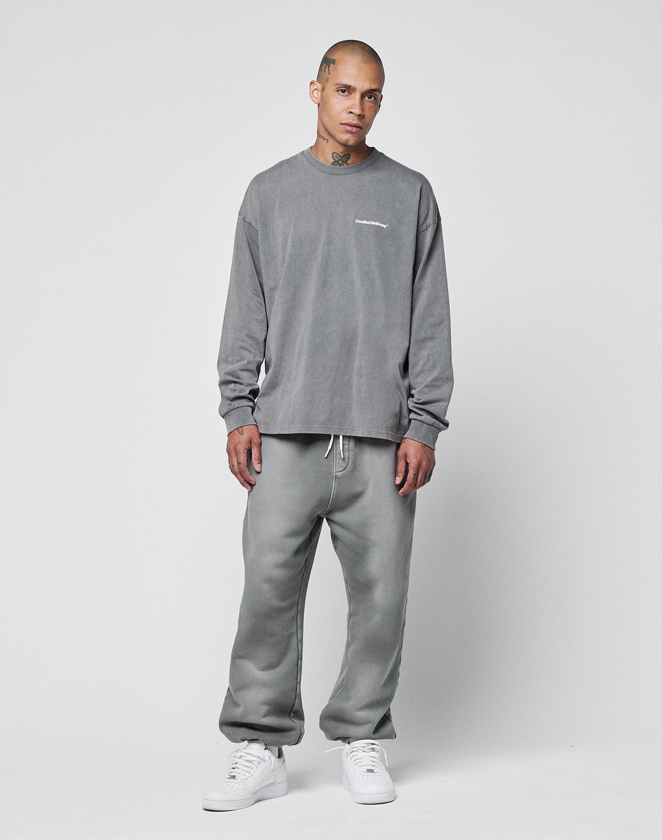 A person with short hair and visible neck and hand tattoos wears LIVE FAST DIE YOUNGās Basic Loose Jogger setāgray, loose-fit, long-sleeve sweatshirt and sweatpantsāpaired with white sneakers. Subtle logo print details the outfit.