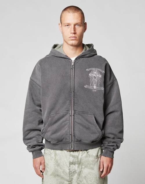 A person with short hair wears the LIVE FAST DIE YOUNG LFDY Cross Hooded Zip in dark gray with a white chest graphic and Waffle Innenfutter, paired with light pants, standing against a plain, light background.