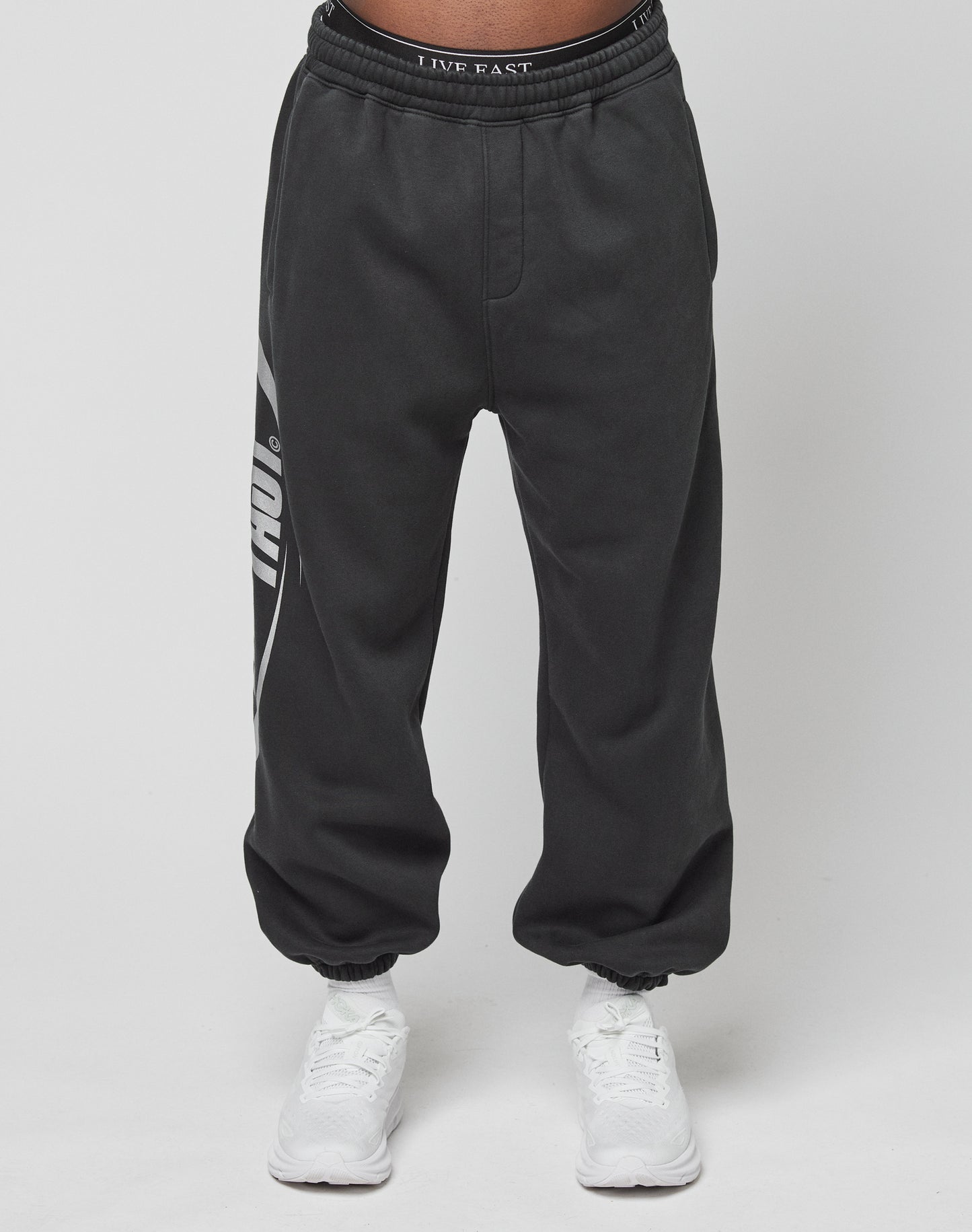A person wears LIVE FAST DIE YOUNG’s LFDY Sphere Jogger, black with a white left-leg graphic and an Elastikbund waistband reading LIVE FAST, paired with white sneakers against a plain white background.