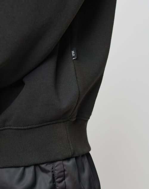 Close-up of a person wearing the LIVE FAST DIE YOUNG Basic Blank Sweater in black, featuring a ribbed waistband made from soft Baumwolle and a small black tag with white text on the side seam.