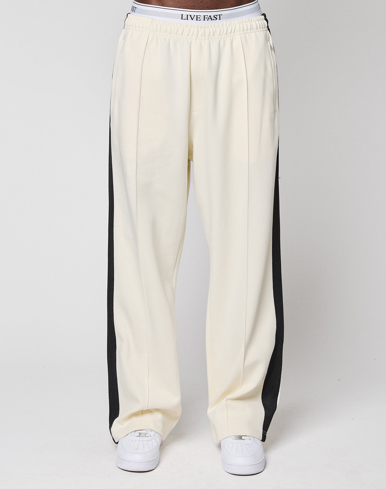 A person wears cream LFDY Striped Jogger by LIVE FAST DIE YOUNG, featuring black side stripes and an elastic waistband with "LIVE FAST." Paired with white sneakers, the waist-down photo is set against a plain background.