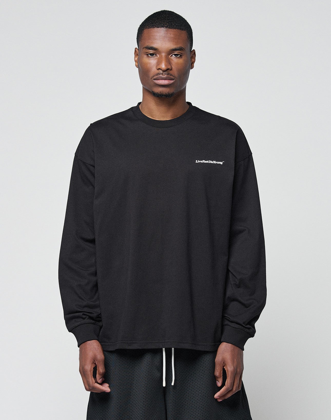 A man faces forward in a LIVE FAST DIE YOUNG Basic Longsleeveāblack, loose-fit, with subtle logo text on the left chestāpaired with black shorts. The oversize style looks relaxed against a plain light gray background.