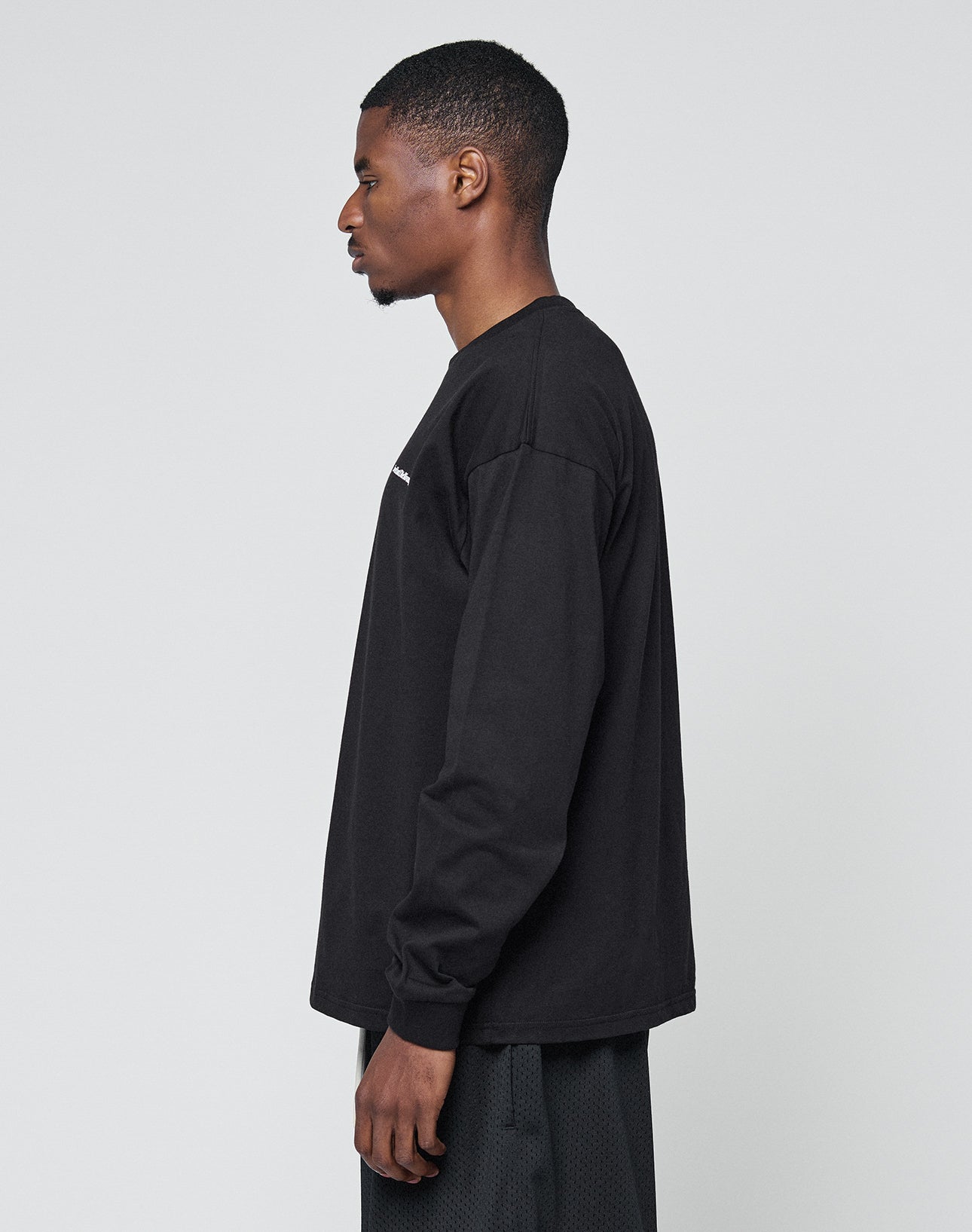 A man in profile faces left, dressed in the LIVE FAST DIE YOUNG Basic Longsleeveā100% Baumwolle black shirtāand oversize-fit black textured pants, against a plain light gray background.