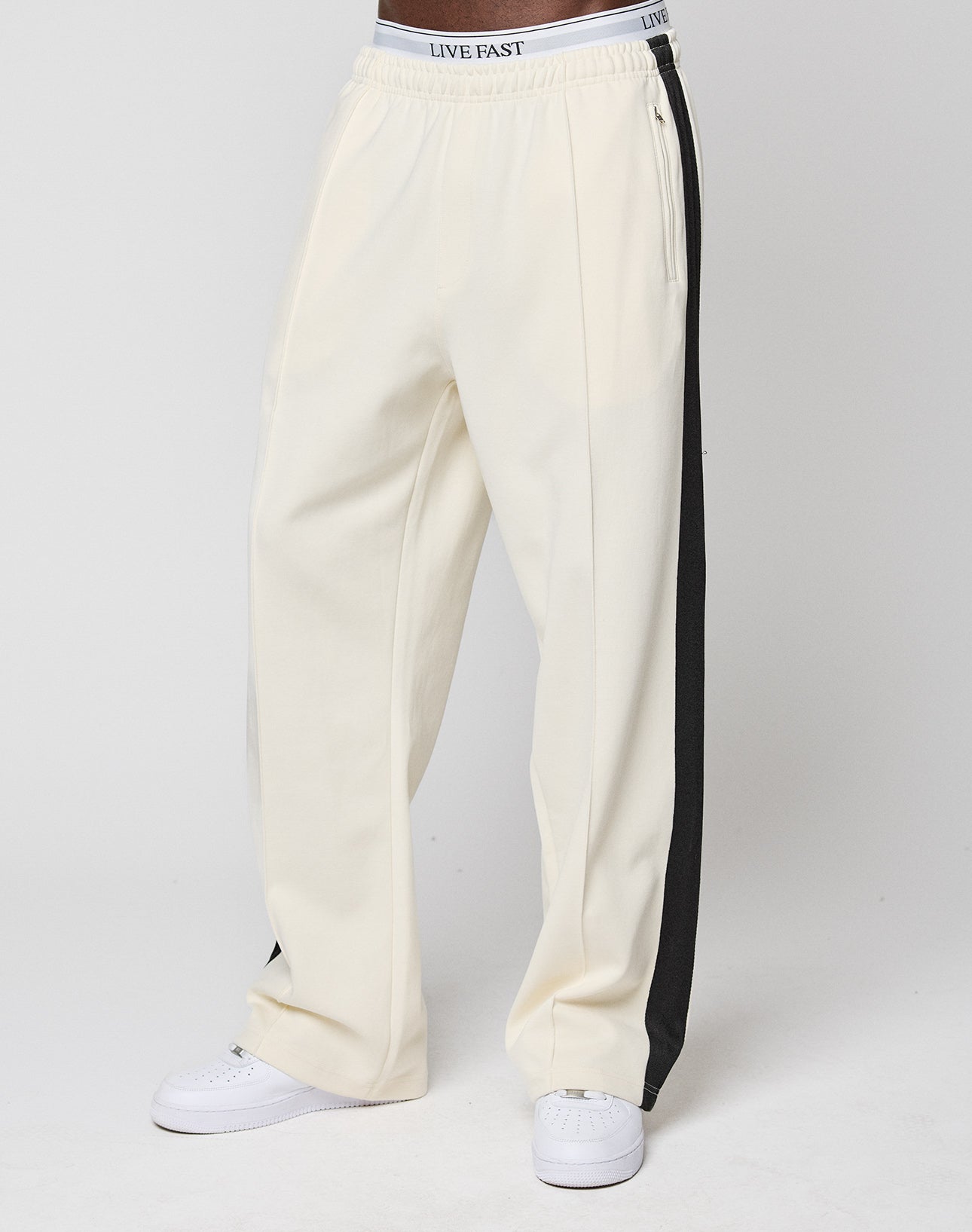 A person wears off-white LFDY Striped Jogger pants by LIVE FAST DIE YOUNG, featuring black side stripes and an elastic waistband with "LIVE FAST" text, paired with white sneakers against a plain white background.