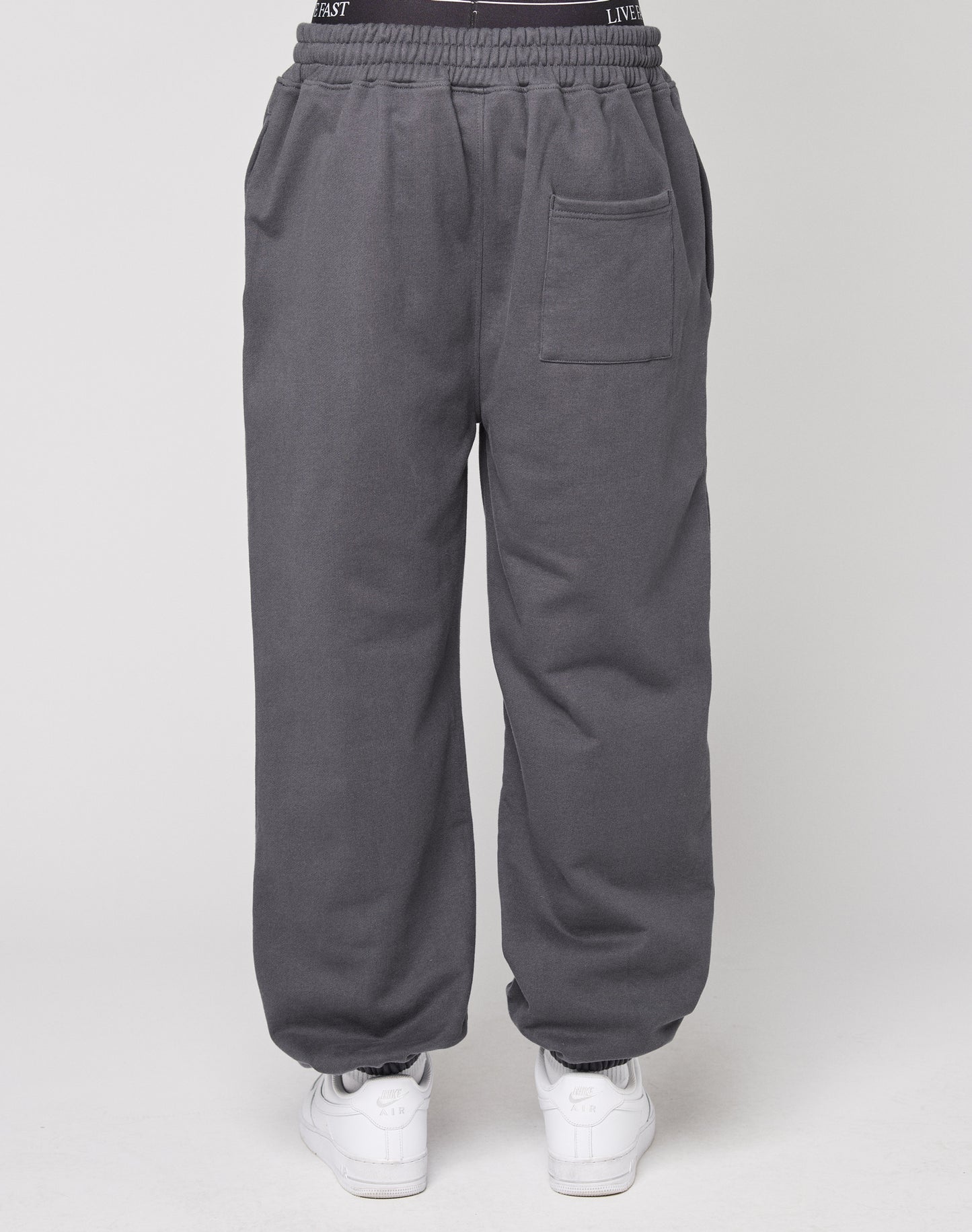Eine Person trƤgt den DY Loose Jogger von LIVE FAST DIE YOUNG in dunkelgrau mit Elastikbund und weiĆen Sneakers, von hinten vor hellem Hintergrund zu sehen.