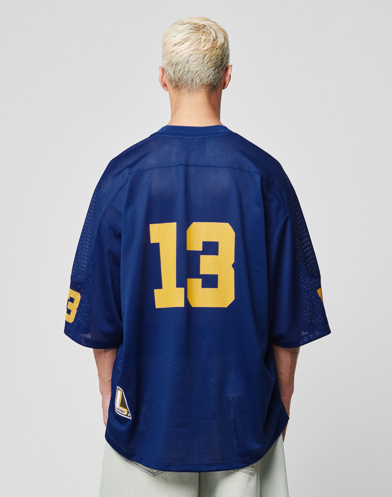A person with short, bleached blond hair faces away, wearing the LIVE FAST DIE YOUNG LFDY Mesh Jersey in Oversize Fit, navy blue with bold yellow number 13 on back and sleeve. The backdrop is plain light gray.