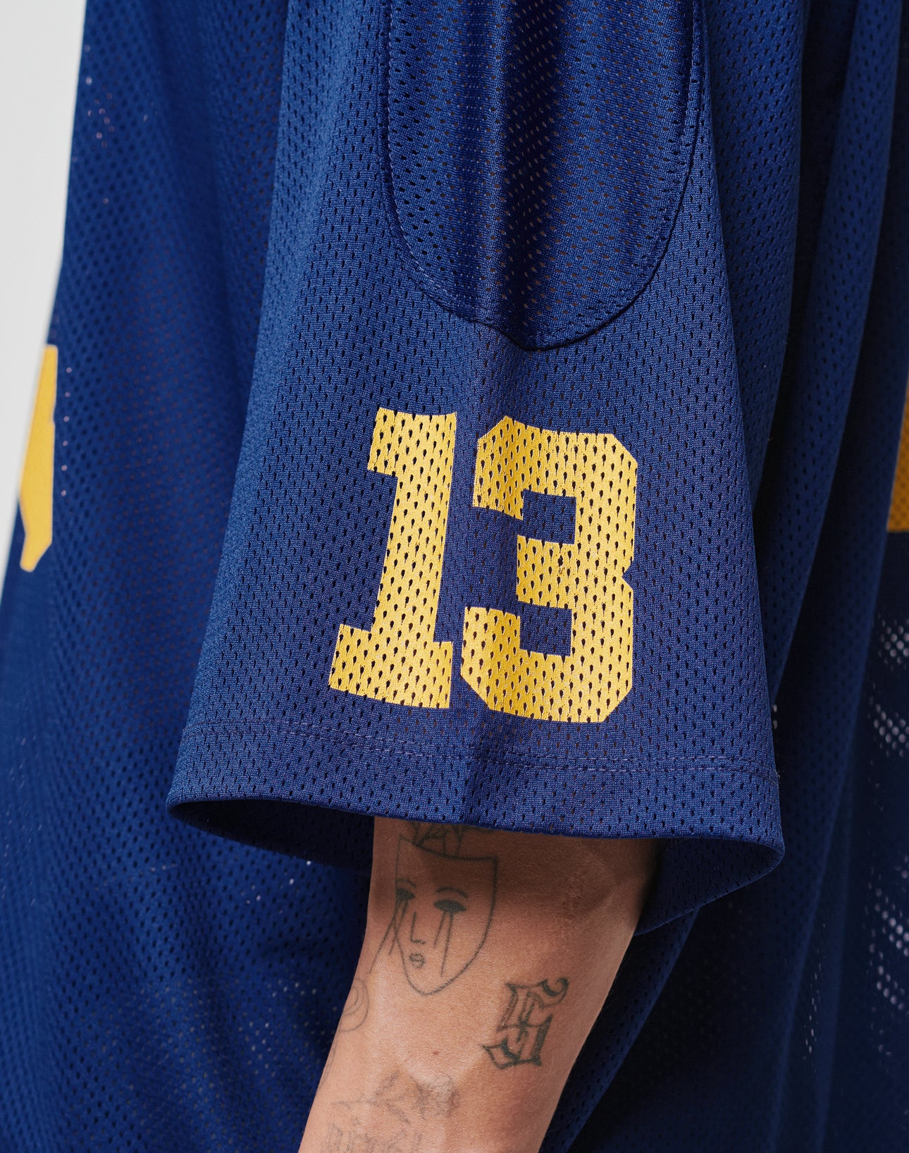 Someone wearing the LFDY Mesh Jersey by LIVE FAST DIE YOUNGâa blue mesh sports top with a yellow number 13 on the sleeve, classic V-Ausschnitt, and Oversize Fitâshows a tattoo of a stylized face on their arm.