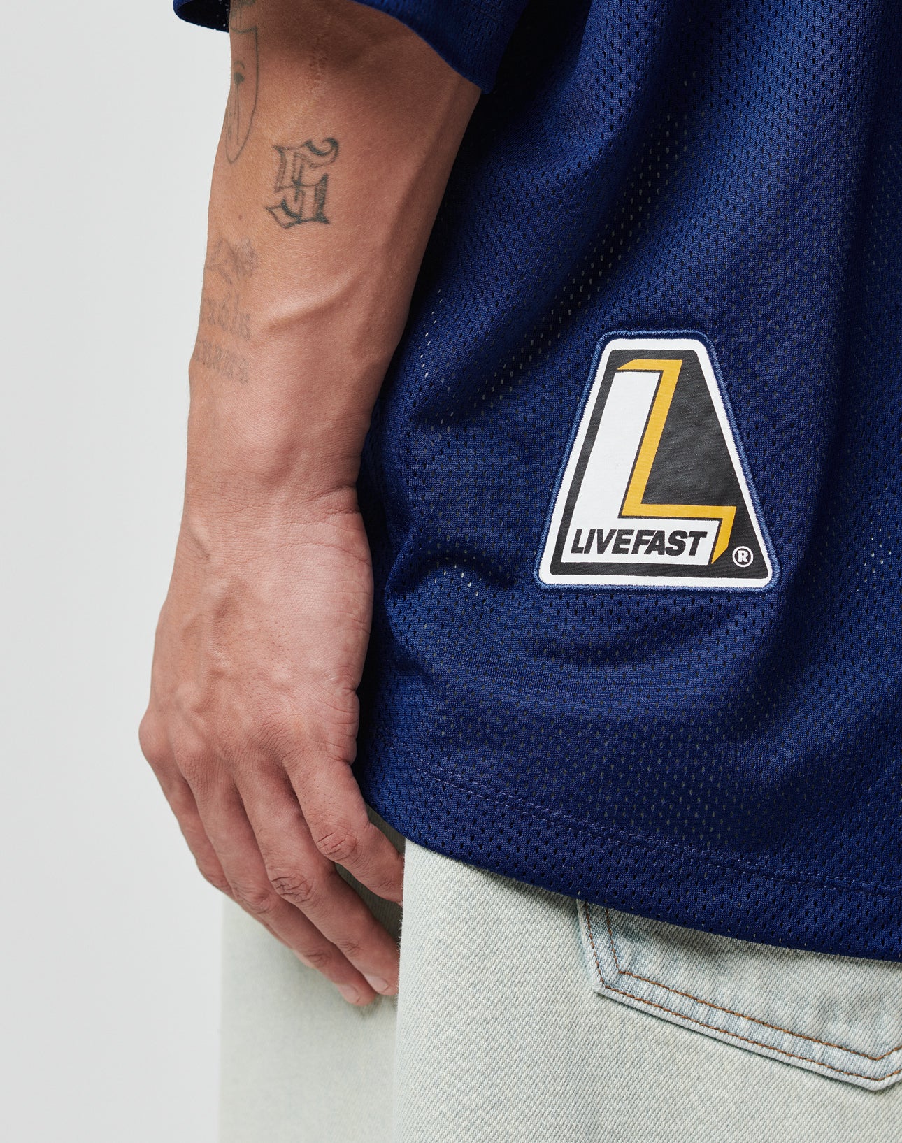 A hand rests beside light-wash jeans, wearing the LIVE FAST DIE YOUNG LFDY Mesh Jersey in navy with an oversize fit and a yellow, black, and white LIVEFAST web patch; initials tattooed on the forearm are also visible.